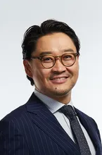 A man wearing glasses, a navy blue pinstripe suit, white shirt, and dark tie, smiling against a white background.