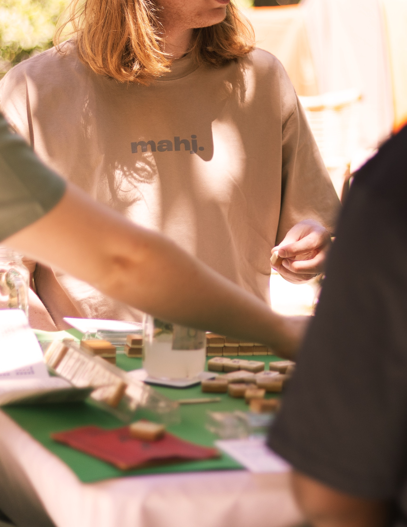 A person with red hair wearing a beige shirt with the word 'mahj.' printed on it, playing Mahjong at a green tabletop with Mahjong tiles and some paper items around.