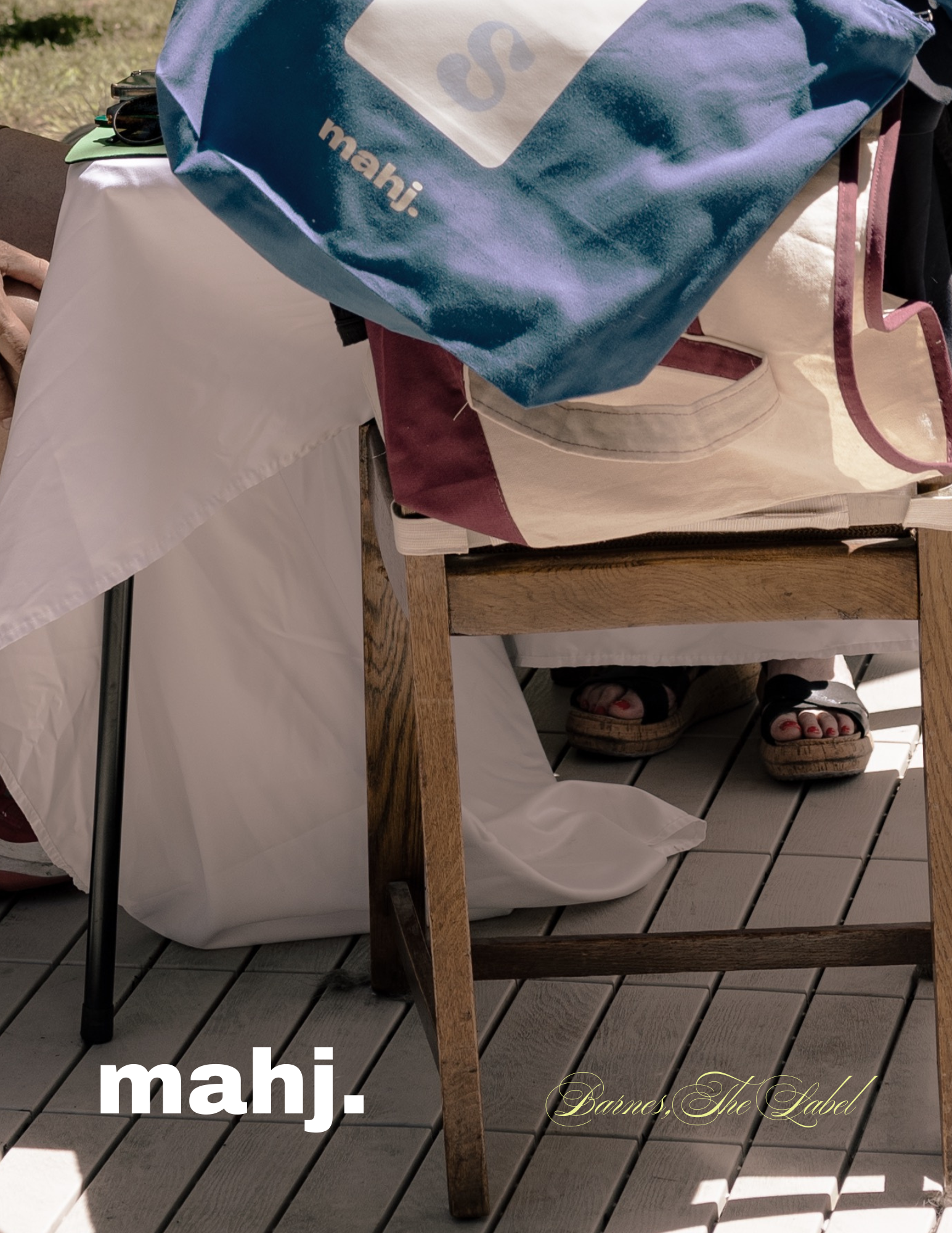 Close-up of a wooden table with a blue bag, a white bag, and a pair of sandals on the outdoor deck, with sunlight and shadows.