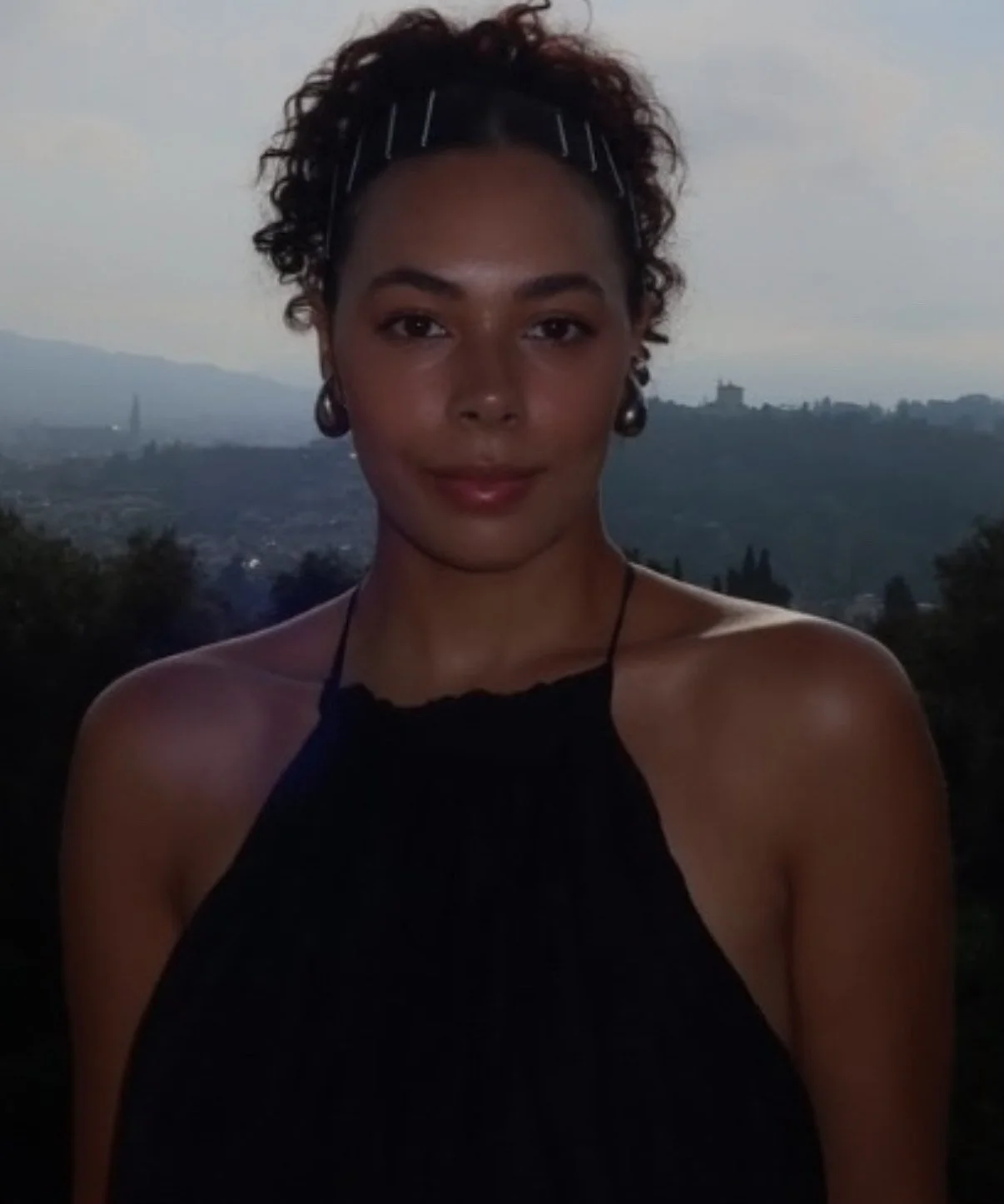 A woman with curly hair, wearing a black halter top, standing outdoors with a landscape of hills and trees in the background during dusk.