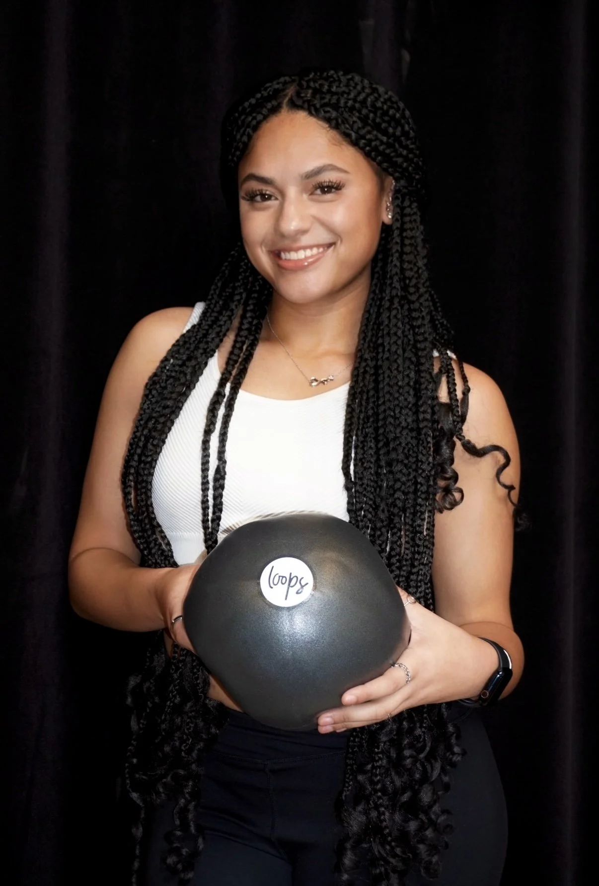 A young woman with long braided hair smiling and holding a black exercise ball labeled 'loop' in front of a dark background.