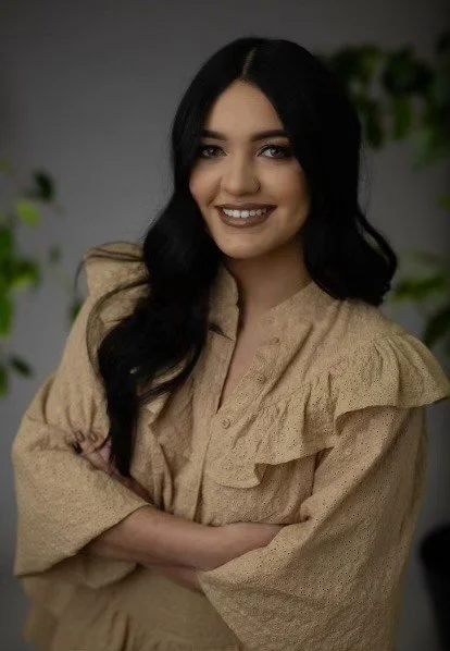 A young woman with long black hair, smiling with arms crossed, wearing a beige blouse with ruffled shoulders, standing in front of a blurred background with some greenery.