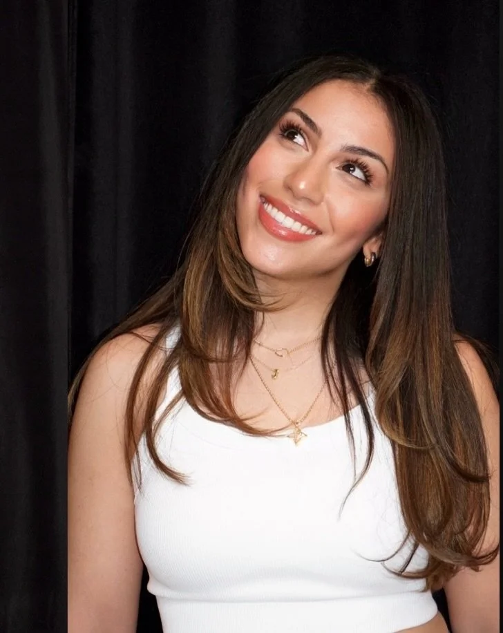 A young woman with brown hair and wearing a white sleeveless top, smiling and looking slightly upwards against a black backdrop.