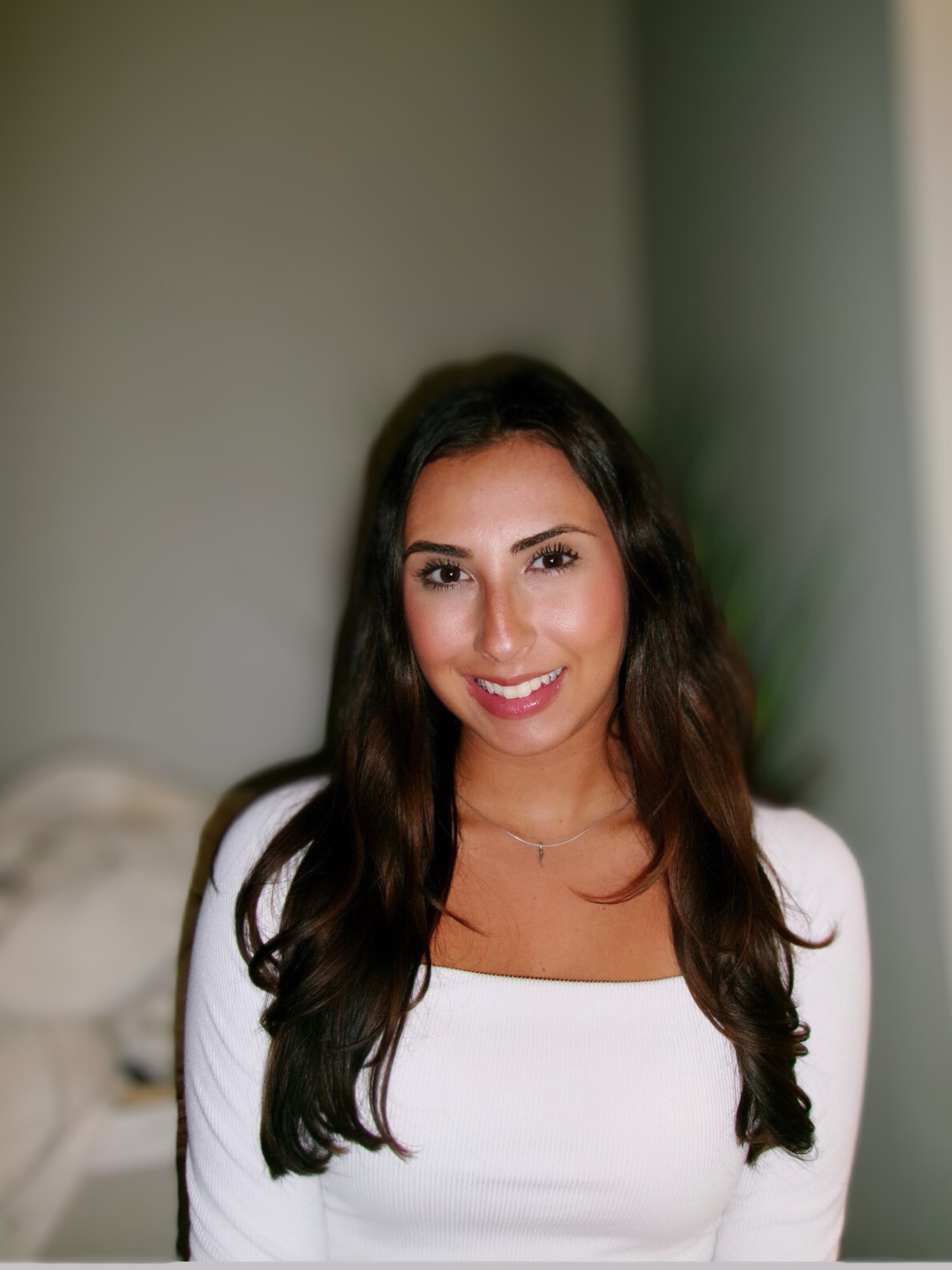 A smiling woman with long dark hair wearing a white top and a silver necklace, indoors with a neutral background.