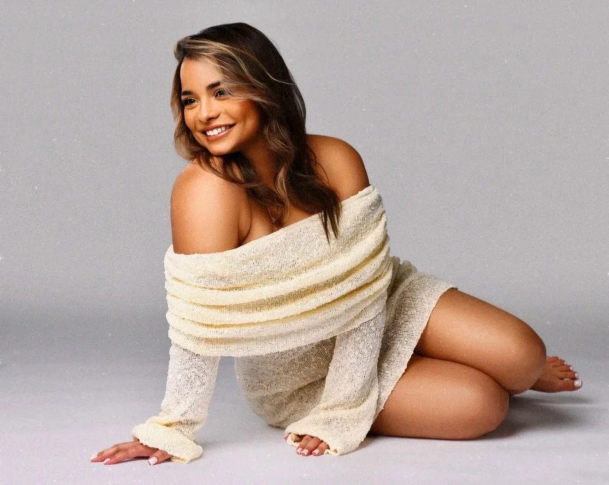 A woman with brown hair sitting on the floor, smiling, wrapped in an off-the-shoulder cream-colored sweater.