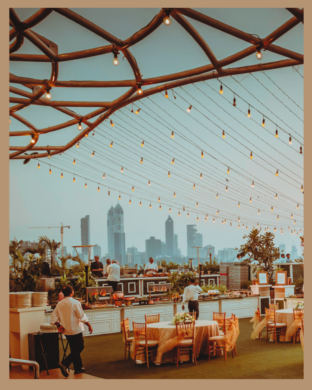 Área de terraza en un restaurante al aire libre con mesas y sillas decoradas con manteles y adornos florales, cocina en continuo proceso con cocineros y personal atendiendo, vista de la ciudad con edificios altos y cielo despejado, iluminación decorativa con guirnaldas de bombillas.