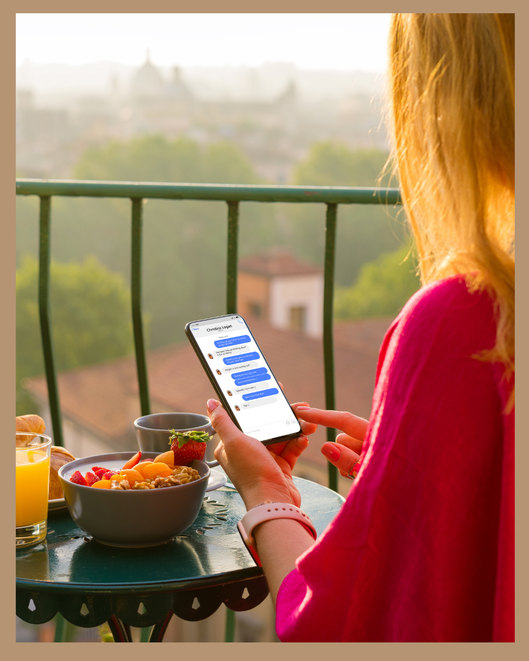 Mujer mirando su teléfono móvil en una terraza con desayuno, includyendo fruta, croissants y jugo, con vista de la ciudad y árboles al atardecer.