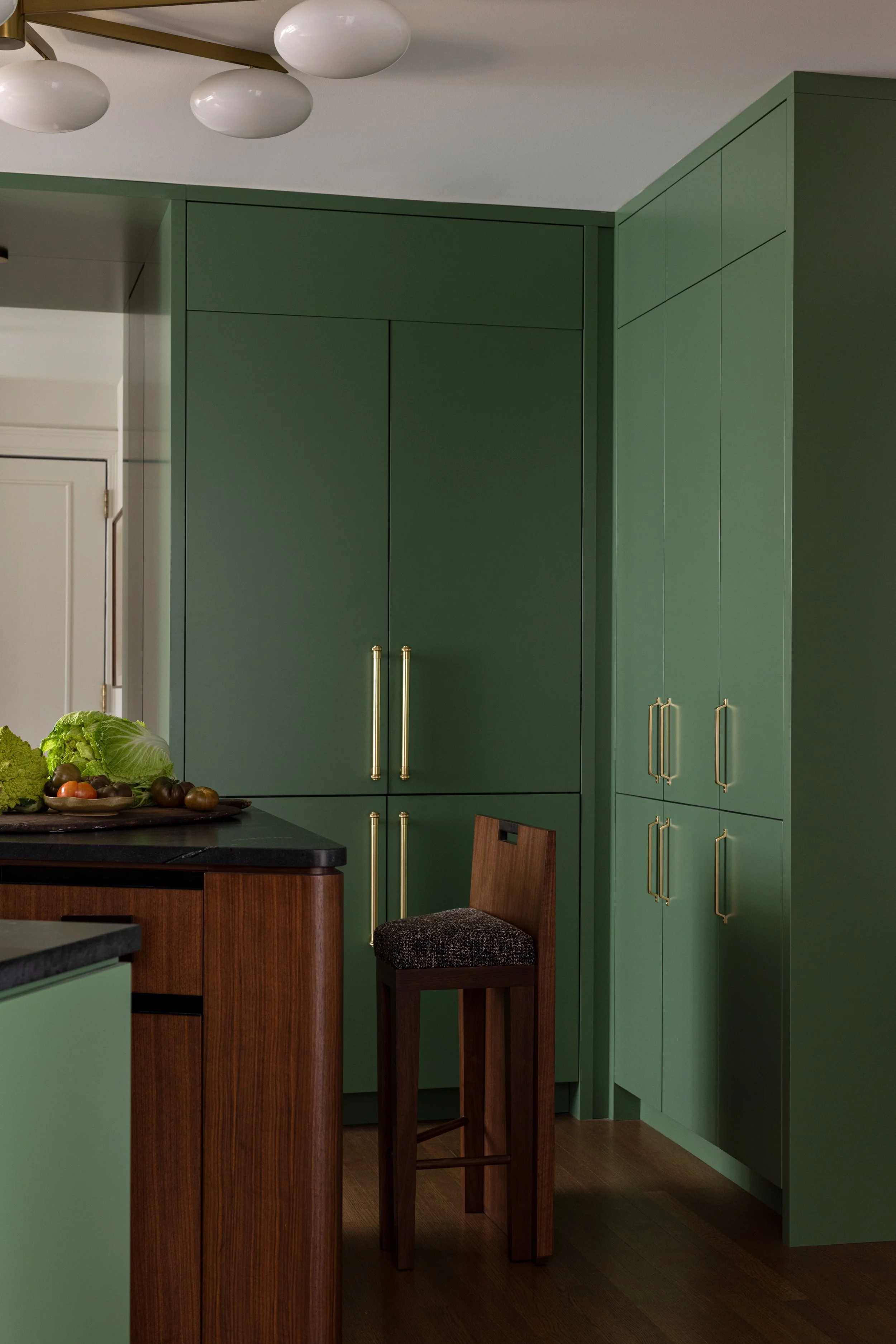 A kitchen corner with green cabinets, a small wooden bar stool with a dark cushion, and a countertop with a lettuce and tomatoes on a tray.