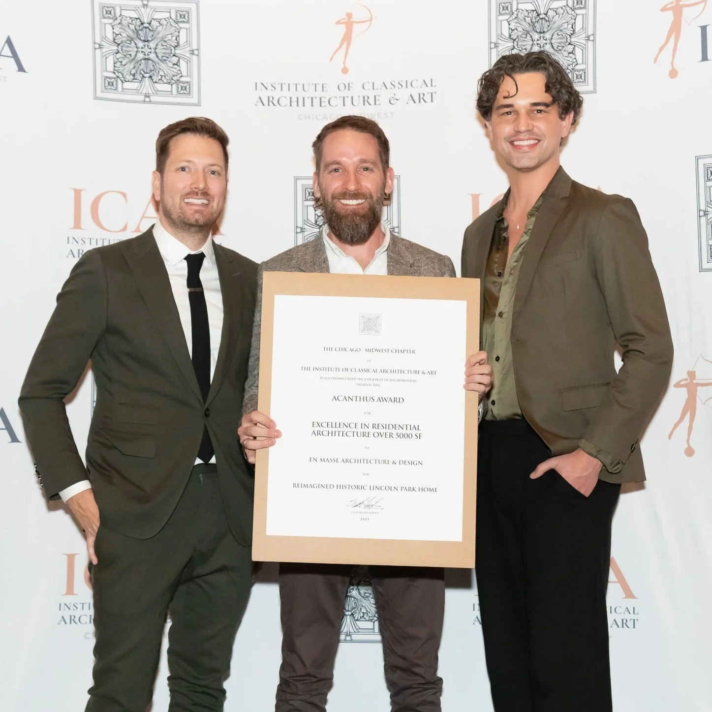 Three men in formal attire standing together, holding a large award certificate at the Institute of Classical Architecture and Art event, with a white backdrop displaying the organization’s logo and name.