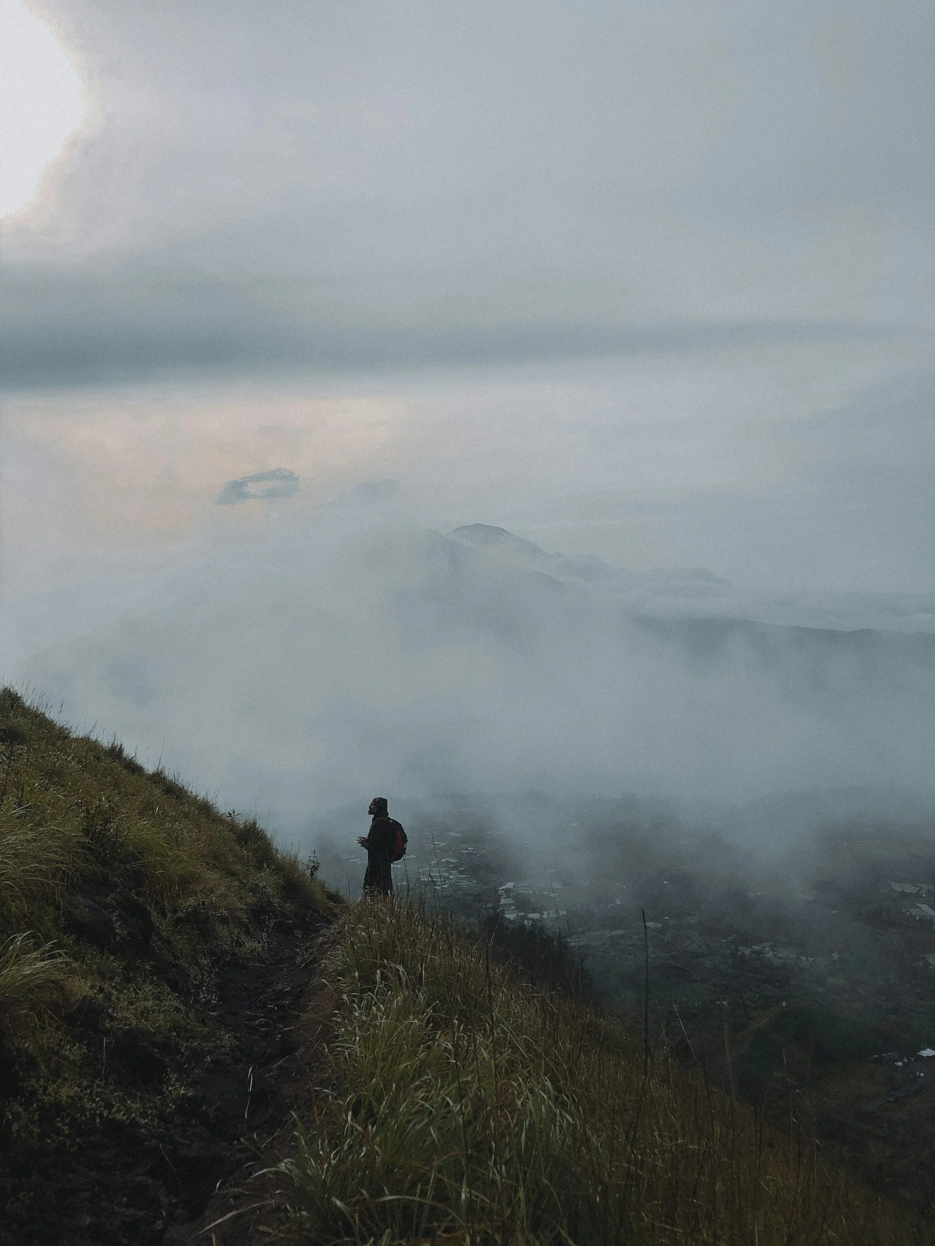 Hiker with a backpack on a grassy mountain trail overlooking a fog-covered valley and distant mountains
