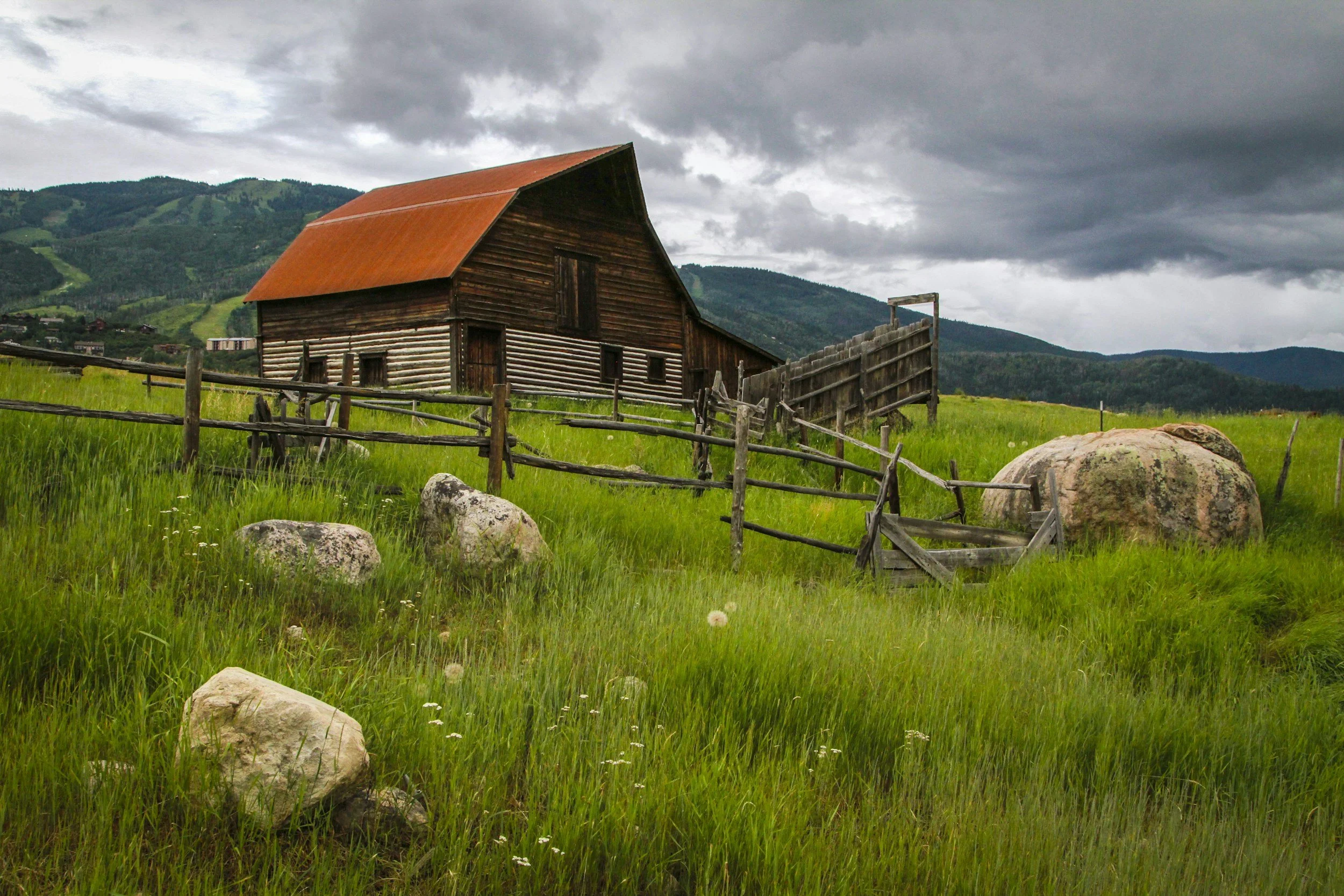 Rustic wooden barn with a red metal roof in a grassy mountain meadow, surrounded by split-rail fences and boulders under stormy clouds
