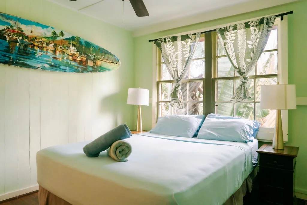 Bedroom with mint green walls, ceiling fan, bed with orange bedding, two windows with patterned curtains, two black nightstands with lamps, and a painting of beach and sea on the wall.