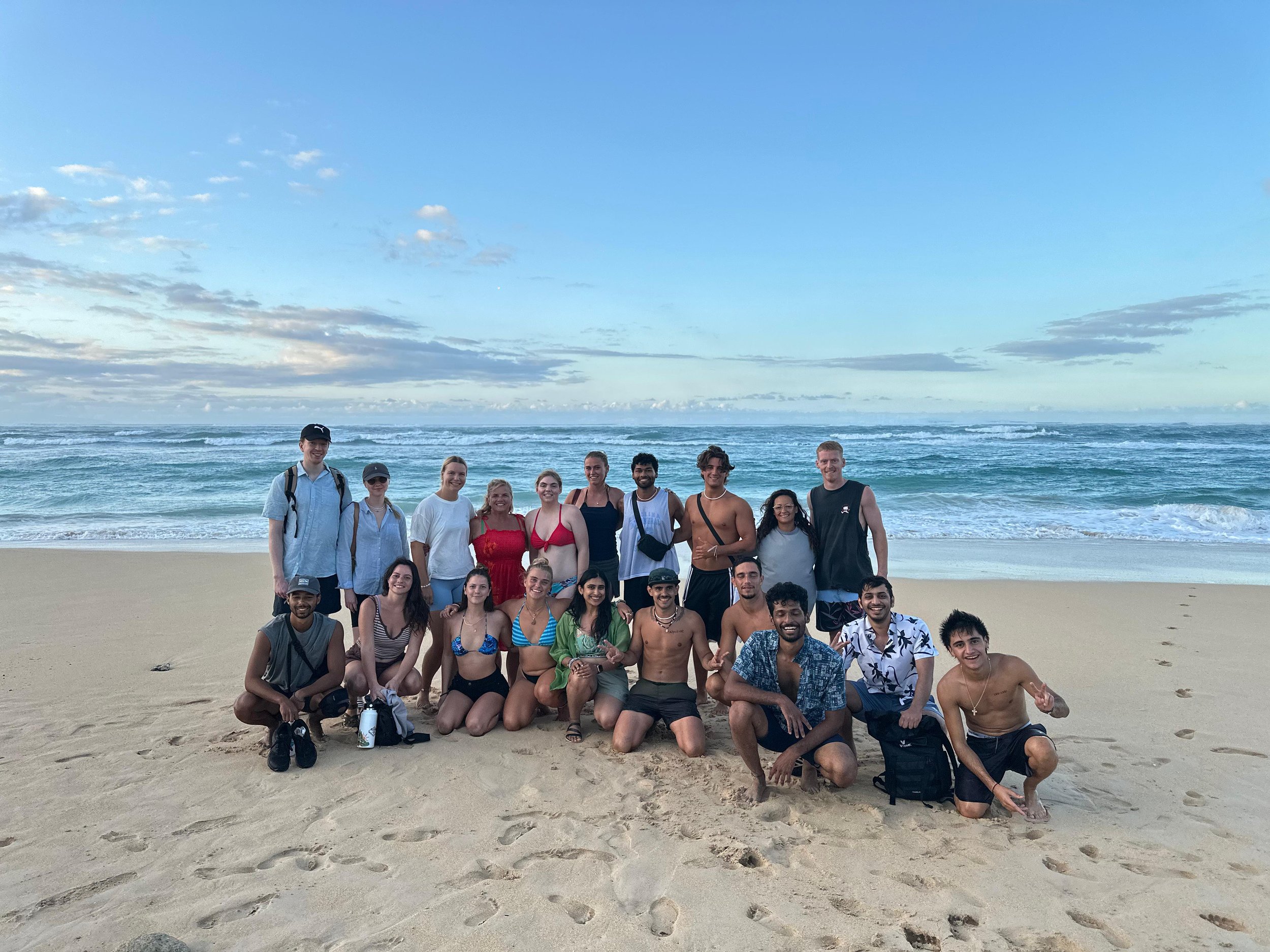 A large group of smiling young people standing and kneeling on a sandy beach with waves and a cloudy sky in the background.