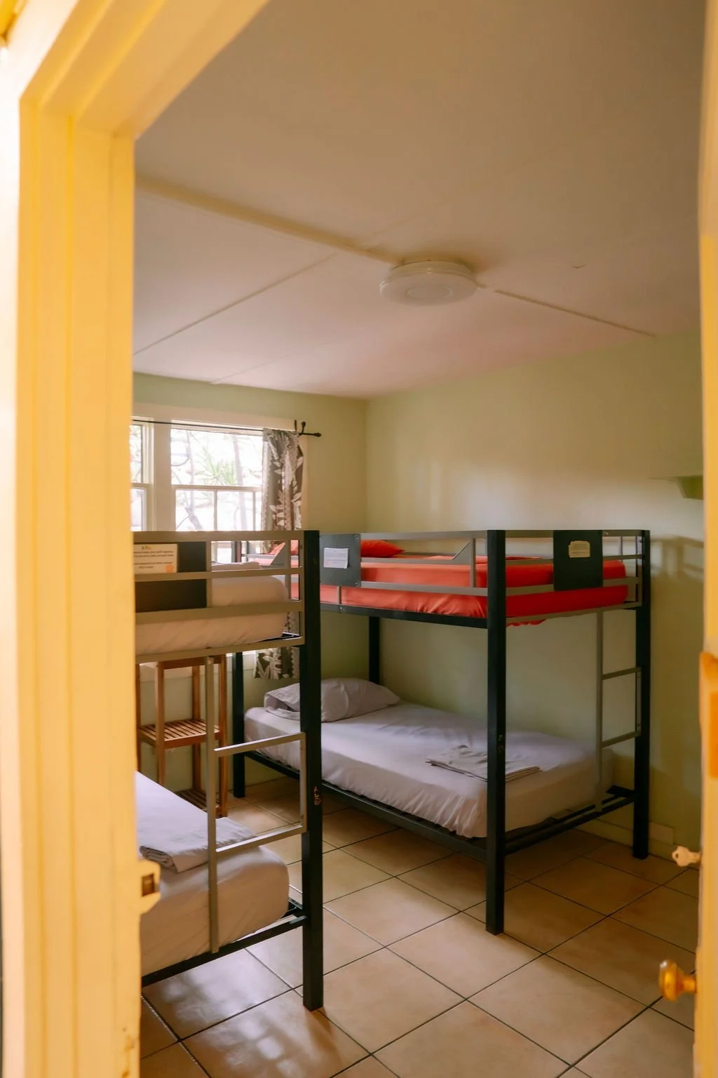 Room with two sets of bunk beds, red bedding, tiled floor, and a window with curtains.