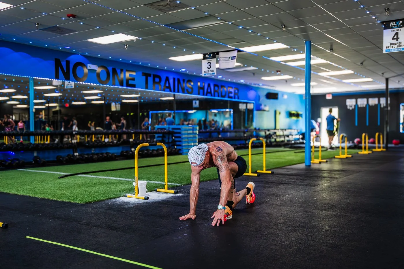 Athlete rising up during burpee broad jumps at NLXF's Hyrox Sim event, with 'No One Trains Harder' quote on the wall, Des Moines