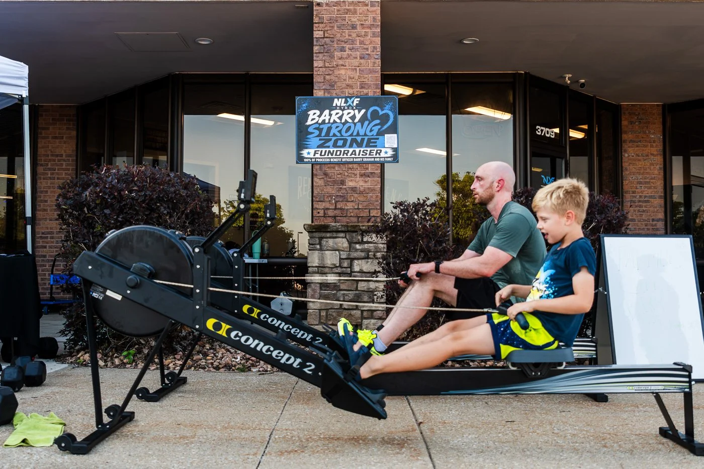 Barry Strong Zone fundraiser sign at NLXF's Hyrox Sim event, with participants on rowing machines in the background, Des Moines