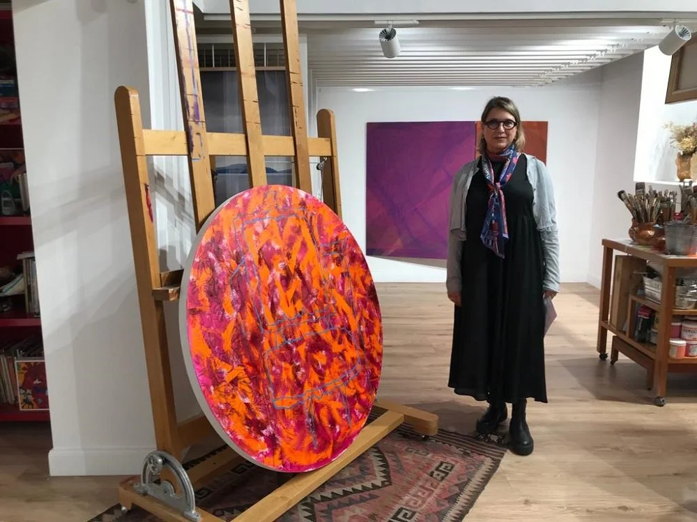 Mujer con gafas y bufanda en una galería de arte, al lado de una obra de arte abstracta en colores naranja, rosado y morado, sobre un caballete.