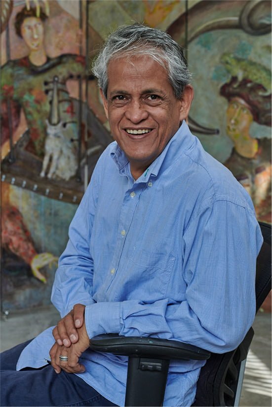 Hombre sonriendo sentado en una silla frente a un mural colorido con figuras humanas y animales.