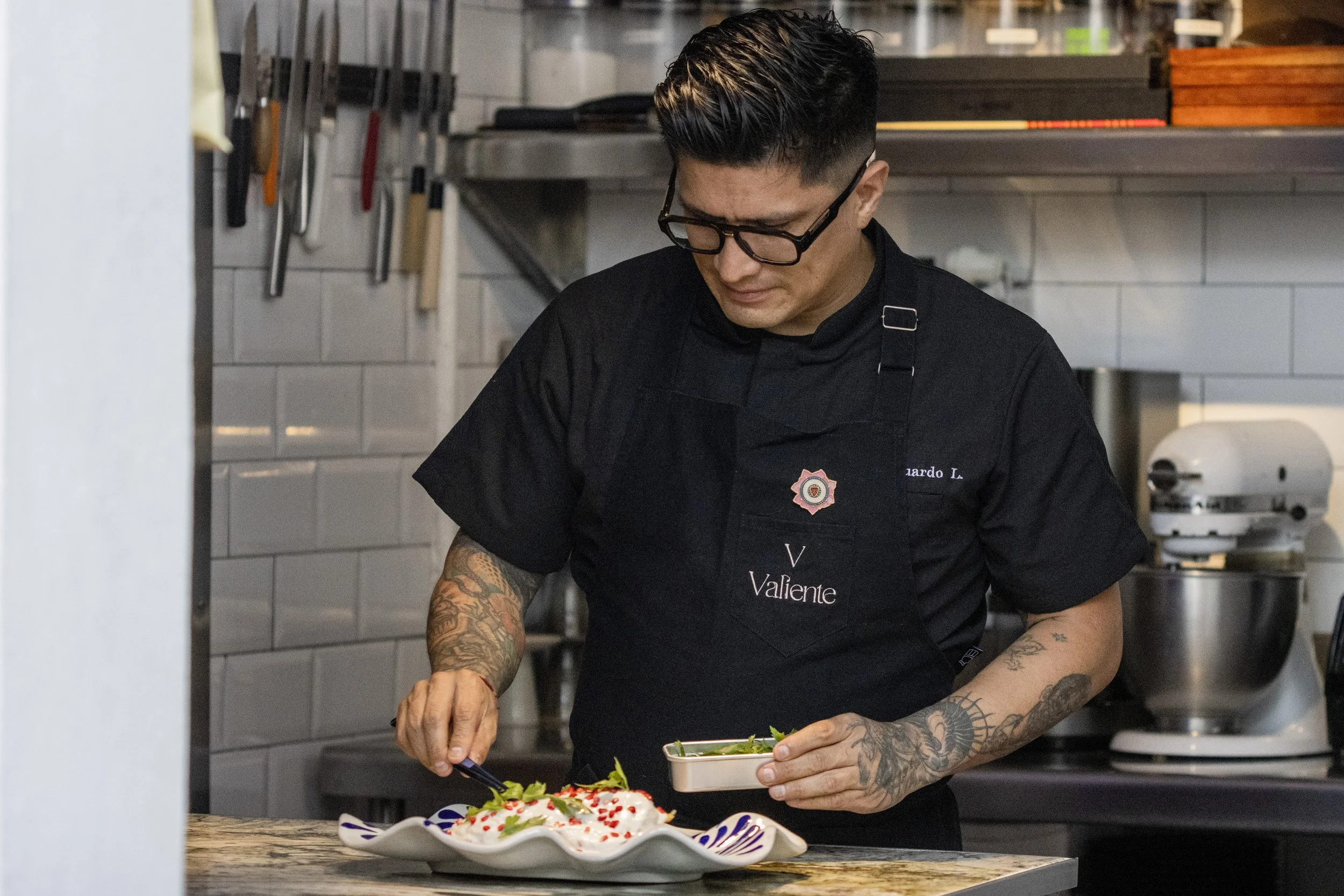 Un chef con gafas y tatuajes decorando un plato en una cocina moderna.