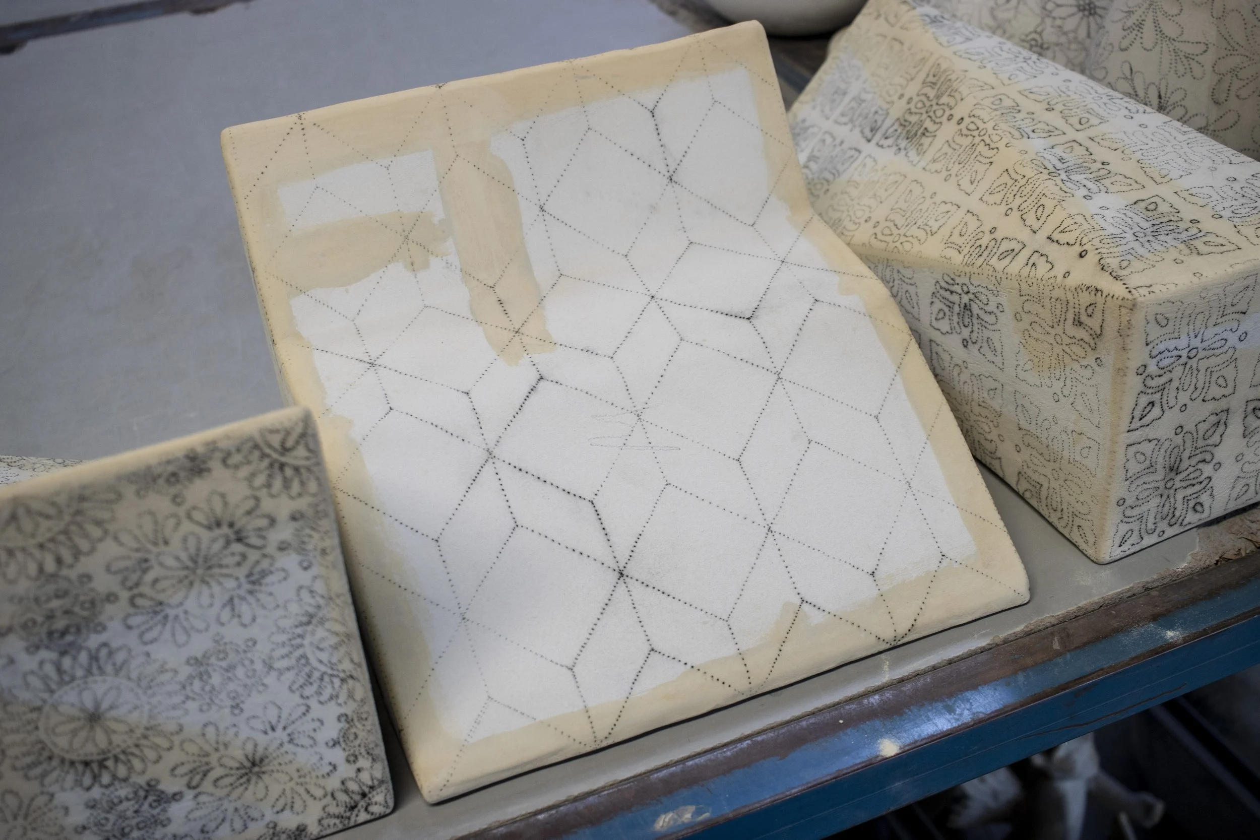 Bloc de cerámica sin esmaltar con patrones de líneas puntiagudas, en proceso de decoración, en una mesa de taller junto a otros bloques con diseños similares.