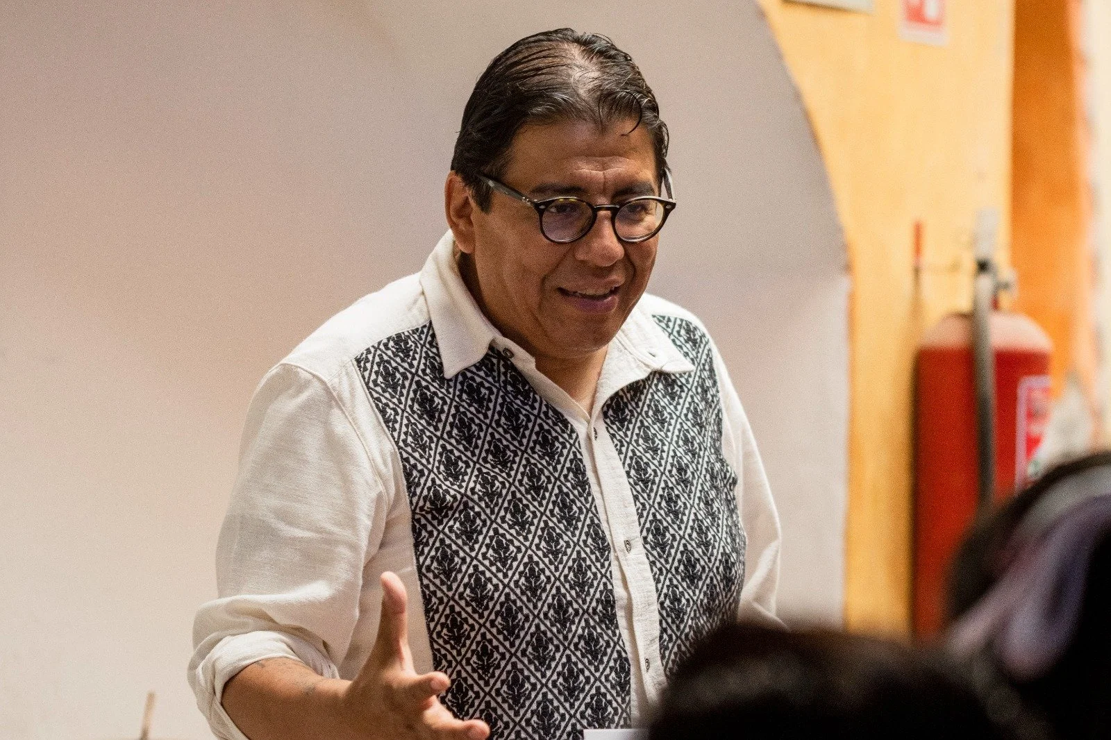 Hombre con gafas y camisa blanca con diseño negro en la parte frontal, hablando con manos en señal explicativa en un ambiente interior.