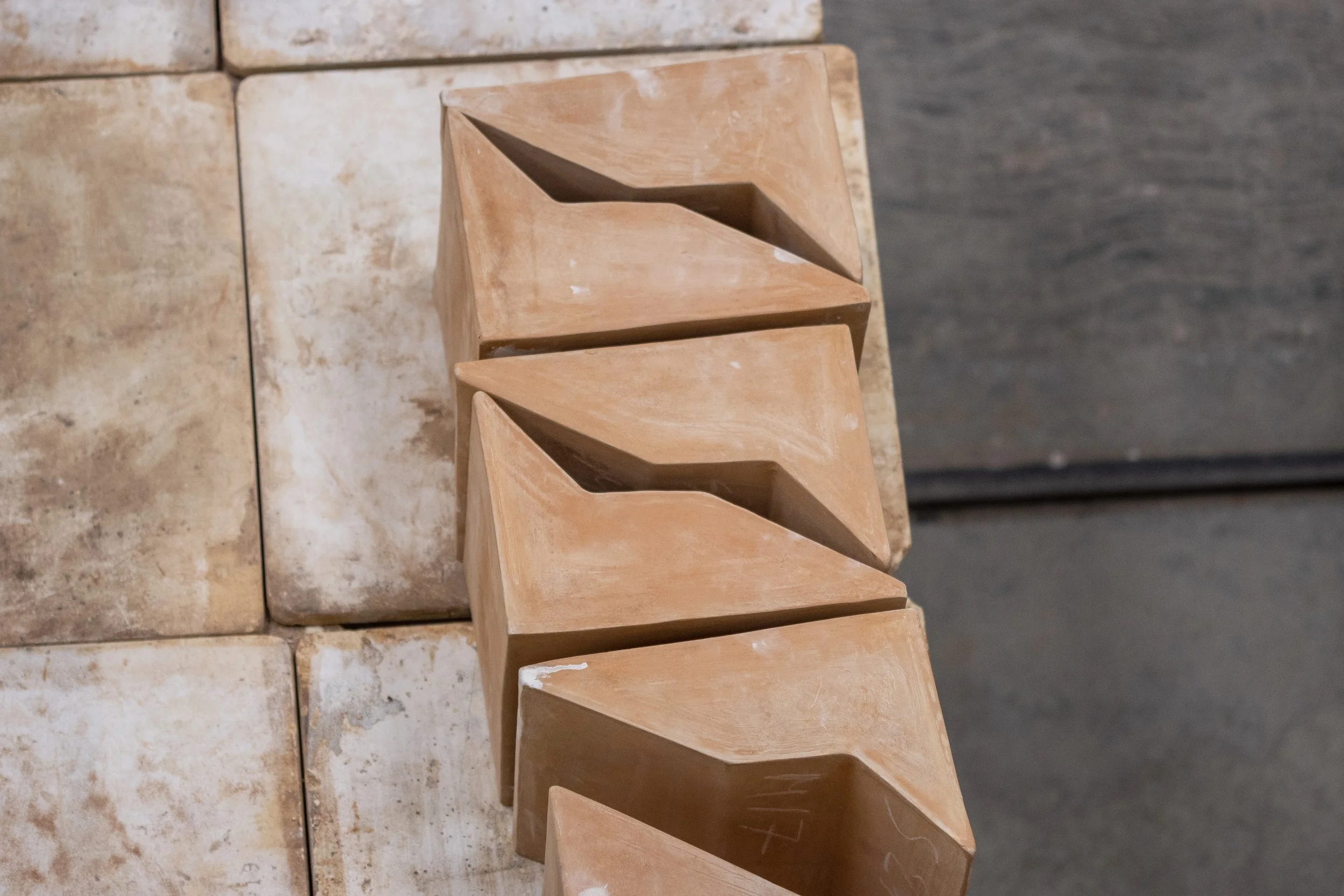 Cuatro bloques de barro cocido con forma triangular alineados sobre una superficie de ladrillos y una superficie de concreto oscuro en el fondo.