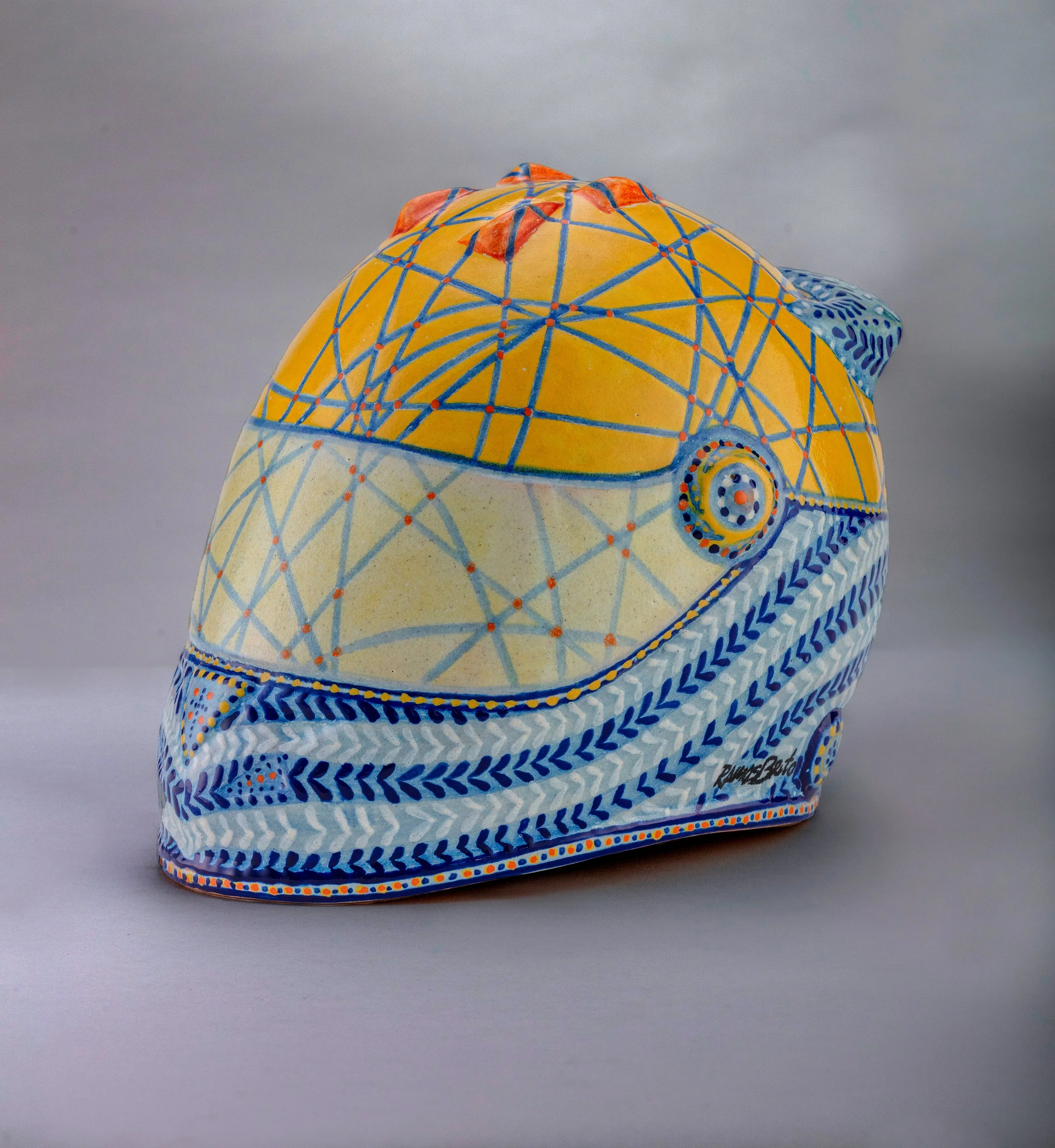 Escultura de un casco de piloto pintado con patrones geométricos en colores amarillo, azul, naranja y blanco.
