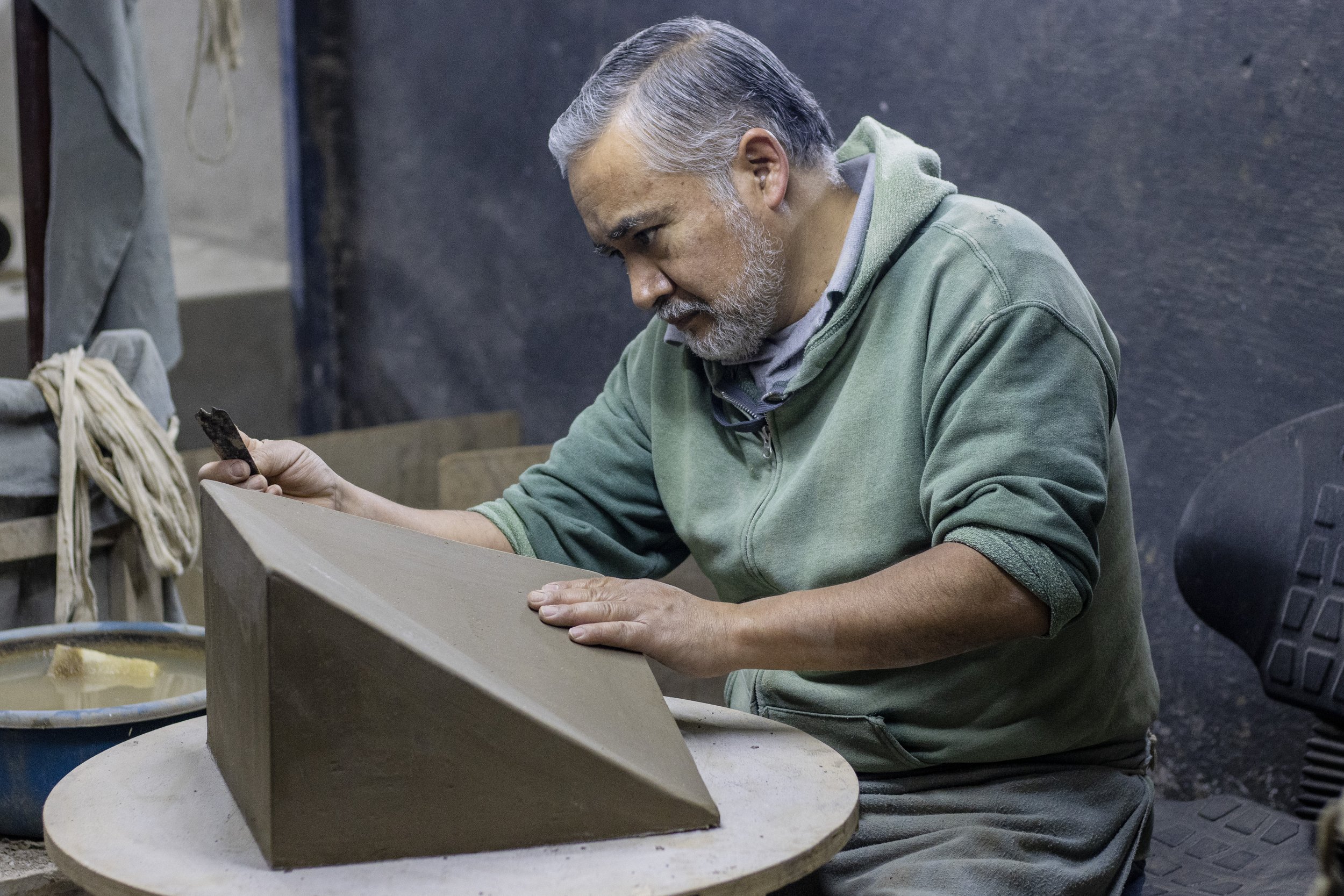 Un hombre mayor con barba y cabello gris pintando una pieza de cerámica en un taller.