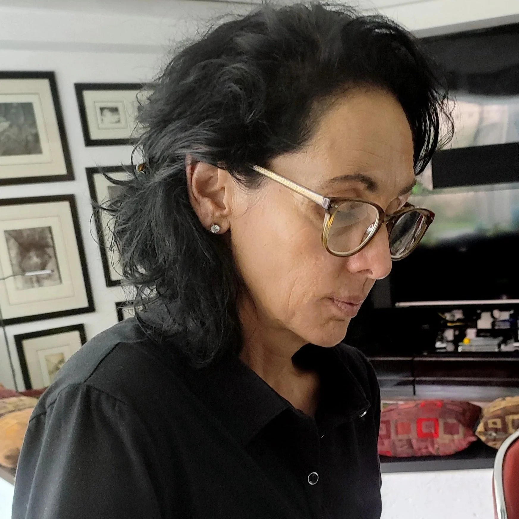 Mujer con cabello oscuro y rizado, usando gafas con marco de carey y pendientes de brillantes, en un entorno interior con paredes decoradas con cuadros, mirando hacia abajo.
