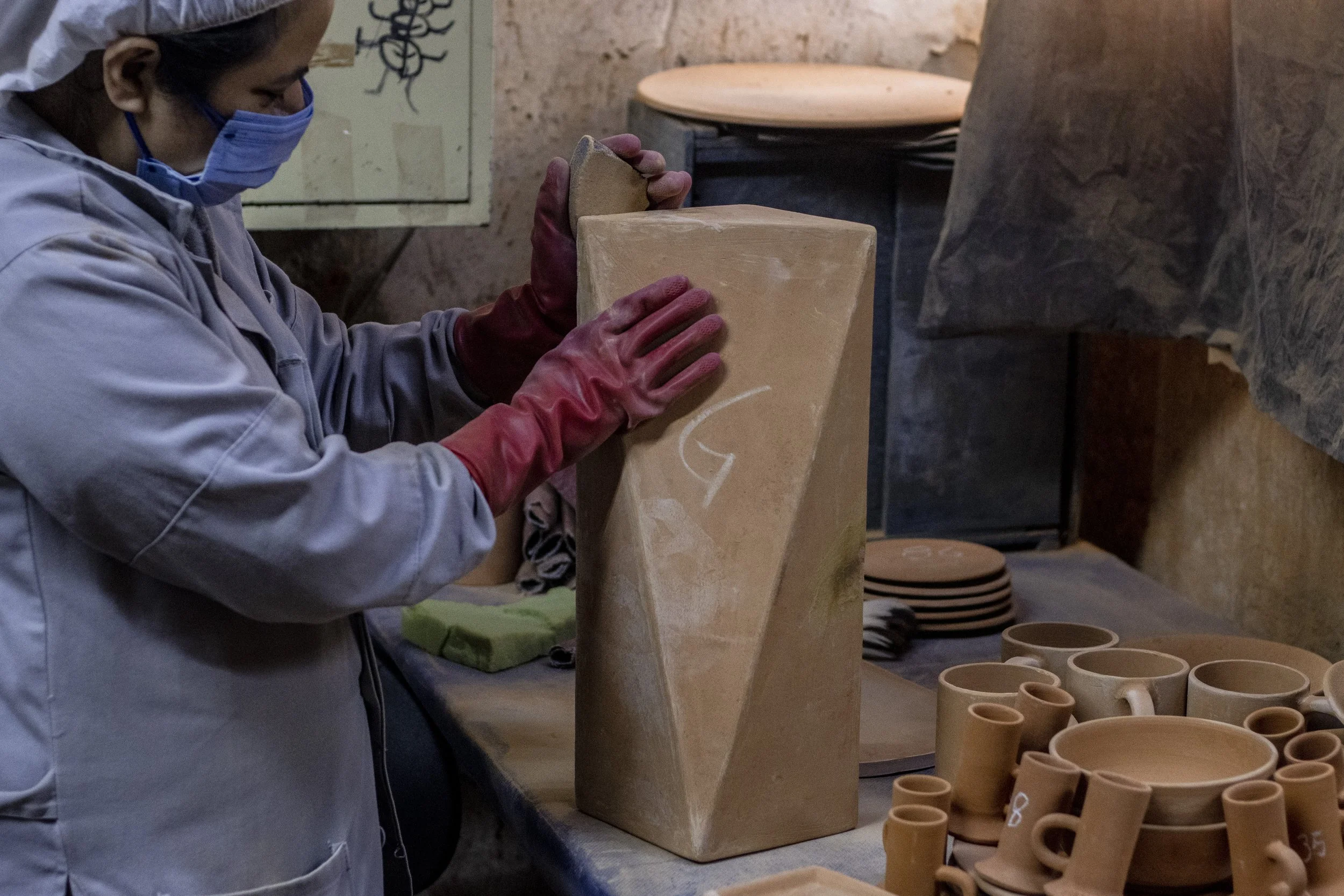 Persona trabajando en cerámica en un taller artesanal, manejando una pieza grande de arcilla o cerámica en forma de cubo con un cincel o herramienta. Rodeada de otros utensilios de cerámica en la mesa.