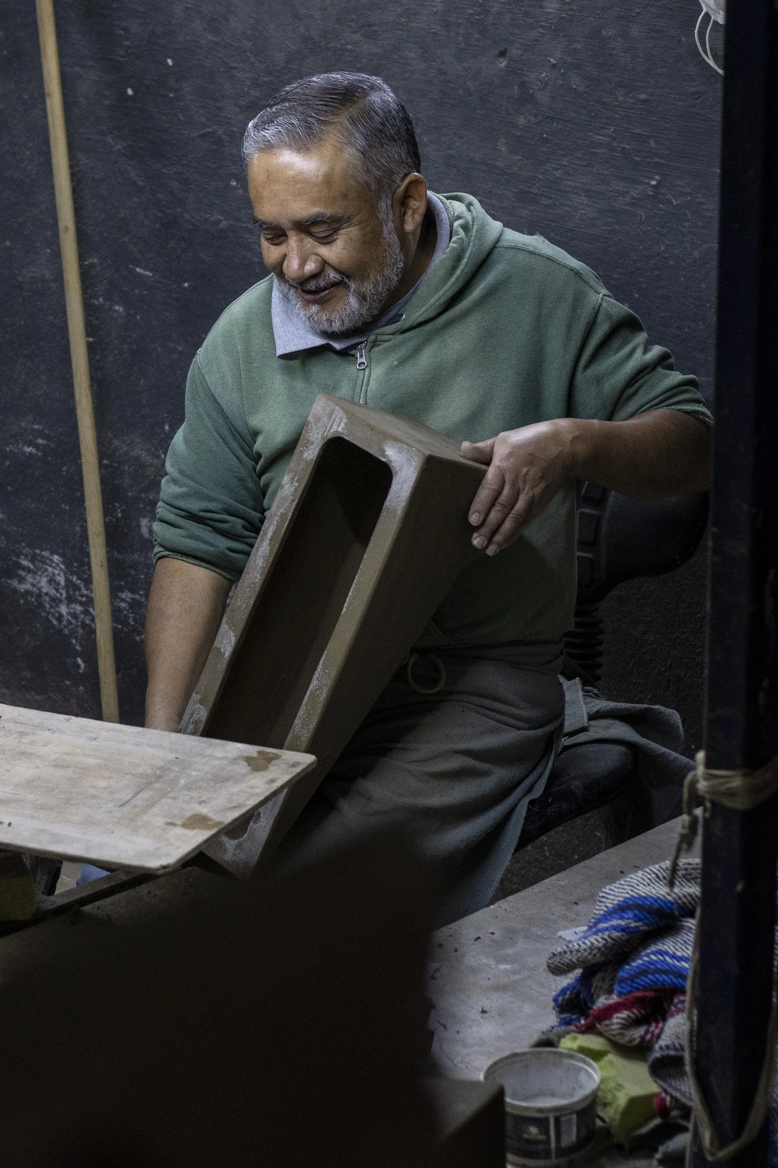 Un hombre mayor trabajando con madera en un taller, sosteniendo un bloque de madera y sonriendo.