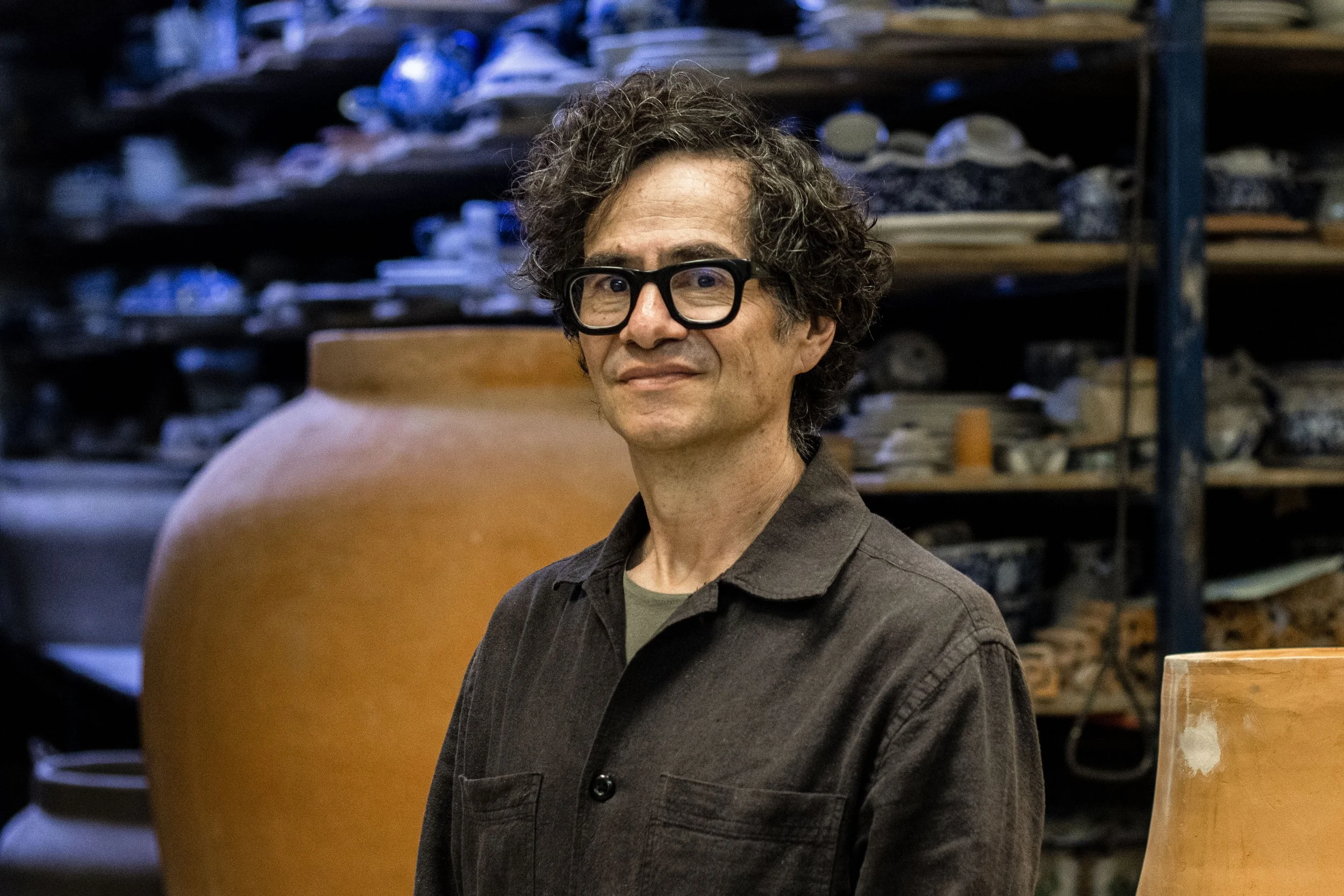 Hombre con cabello rizado y gafas negras en taller de cerámica, rodeado de vasijas de barro y utensilios.