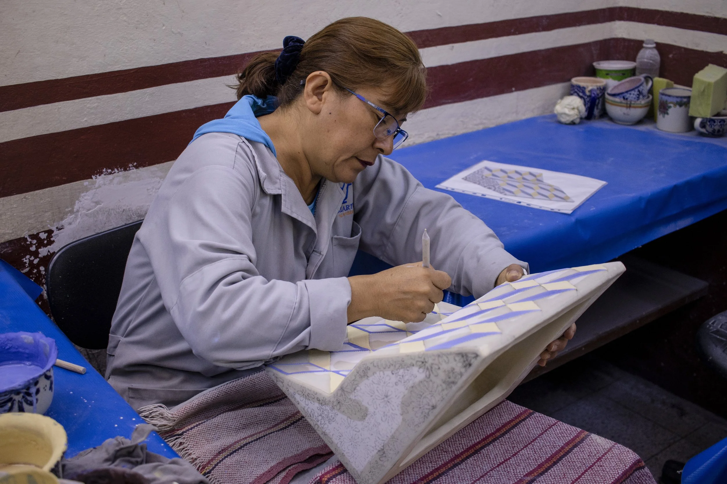 Mujer trabajando en la pintura de cerámica, sentada en una mesa cubierta con tela y rodeada de recipientes y utensilios.