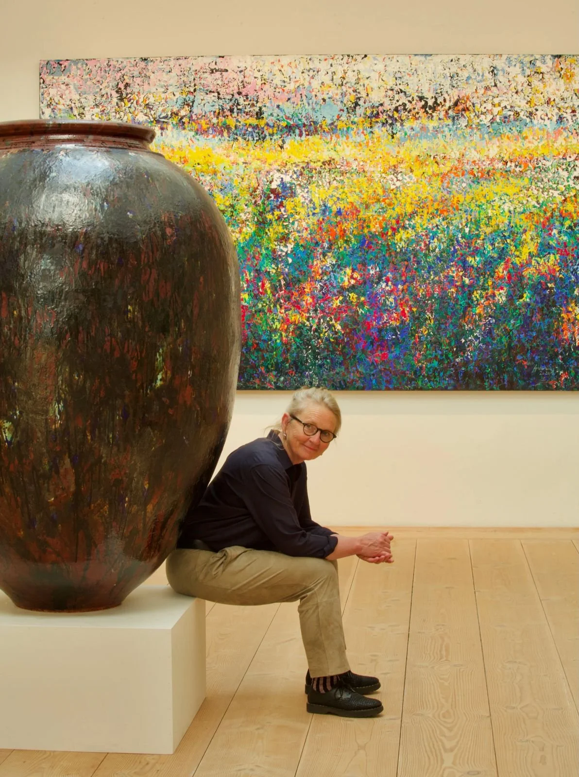 Una mujer con gafas y cabello gris, vestida con camisa azul y pantalones beige, sentada junto a una gran vasija de cerámica en un museo de arte, con una pintura abstracta colorida en el fondo.