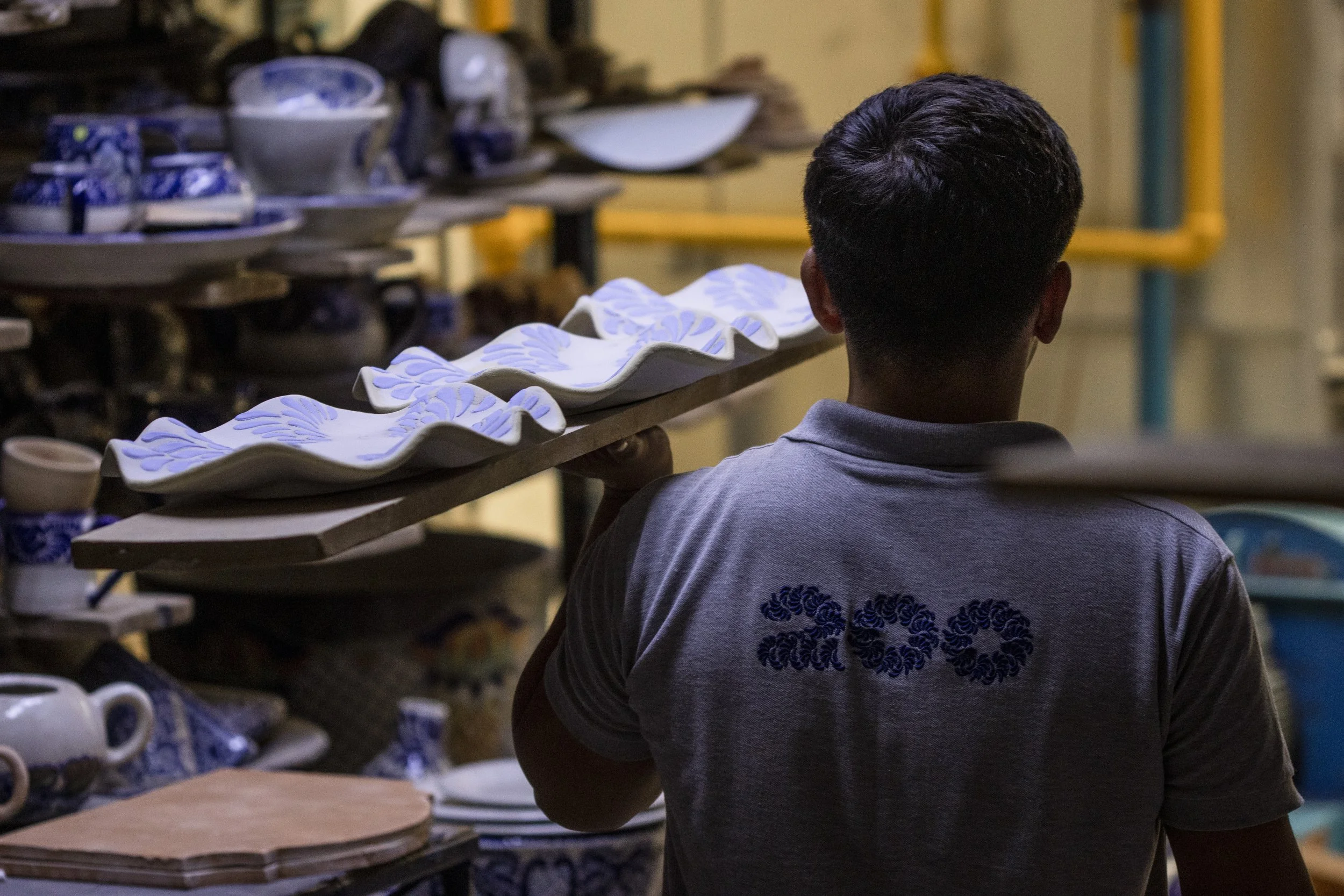 Un hombre con camiseta gris sosteniendo una bandeja con platos de cerámica pintados en azul, en un taller de cerámica, rodeado de más cerámicas y utensilios.