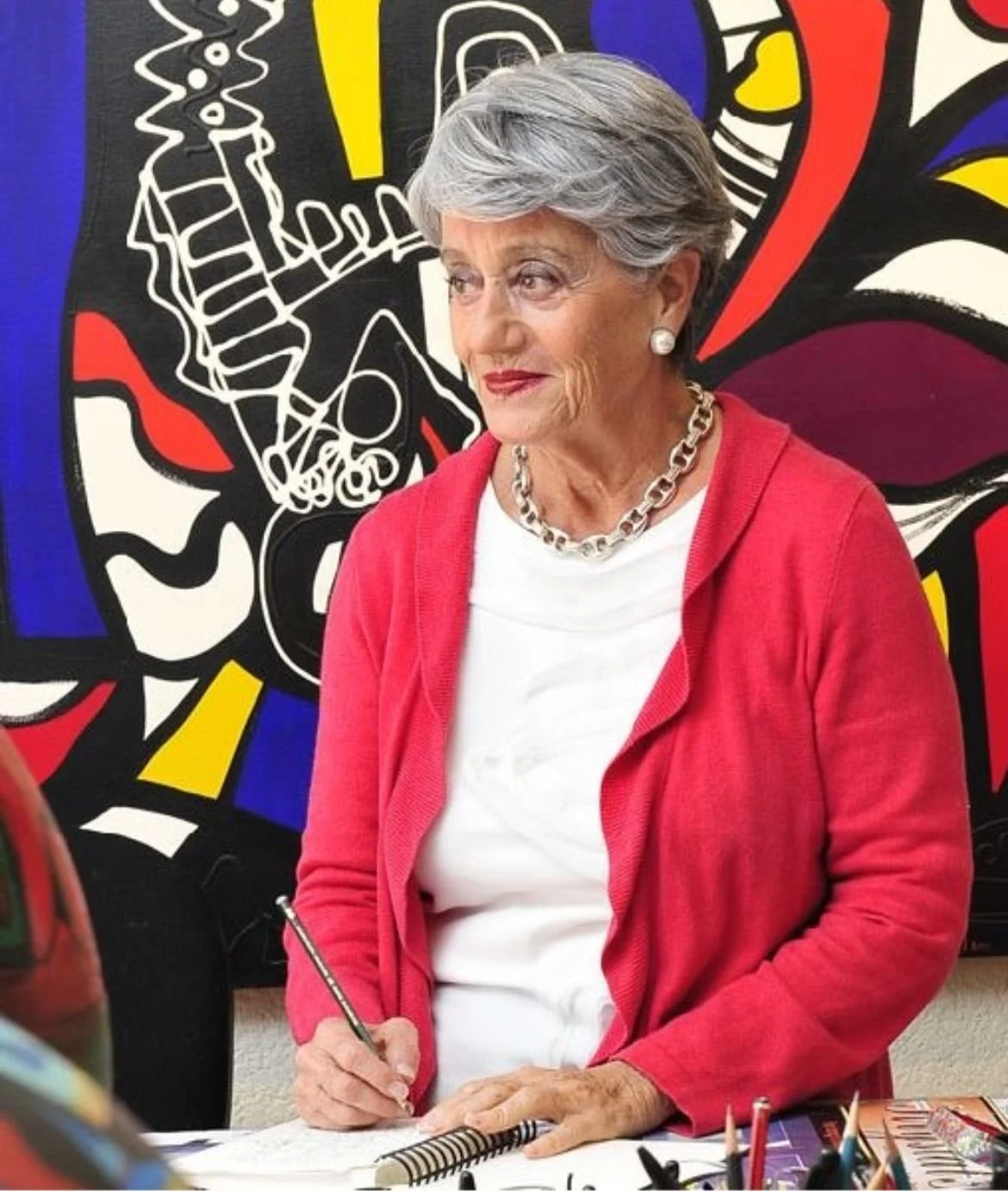 Una mujer de cabello gris y vestido blanco con suéter rojo, usando un collar de cadena, está escribiendo en un cuaderno en una mesa, con una pintura abstracta en el fondo.