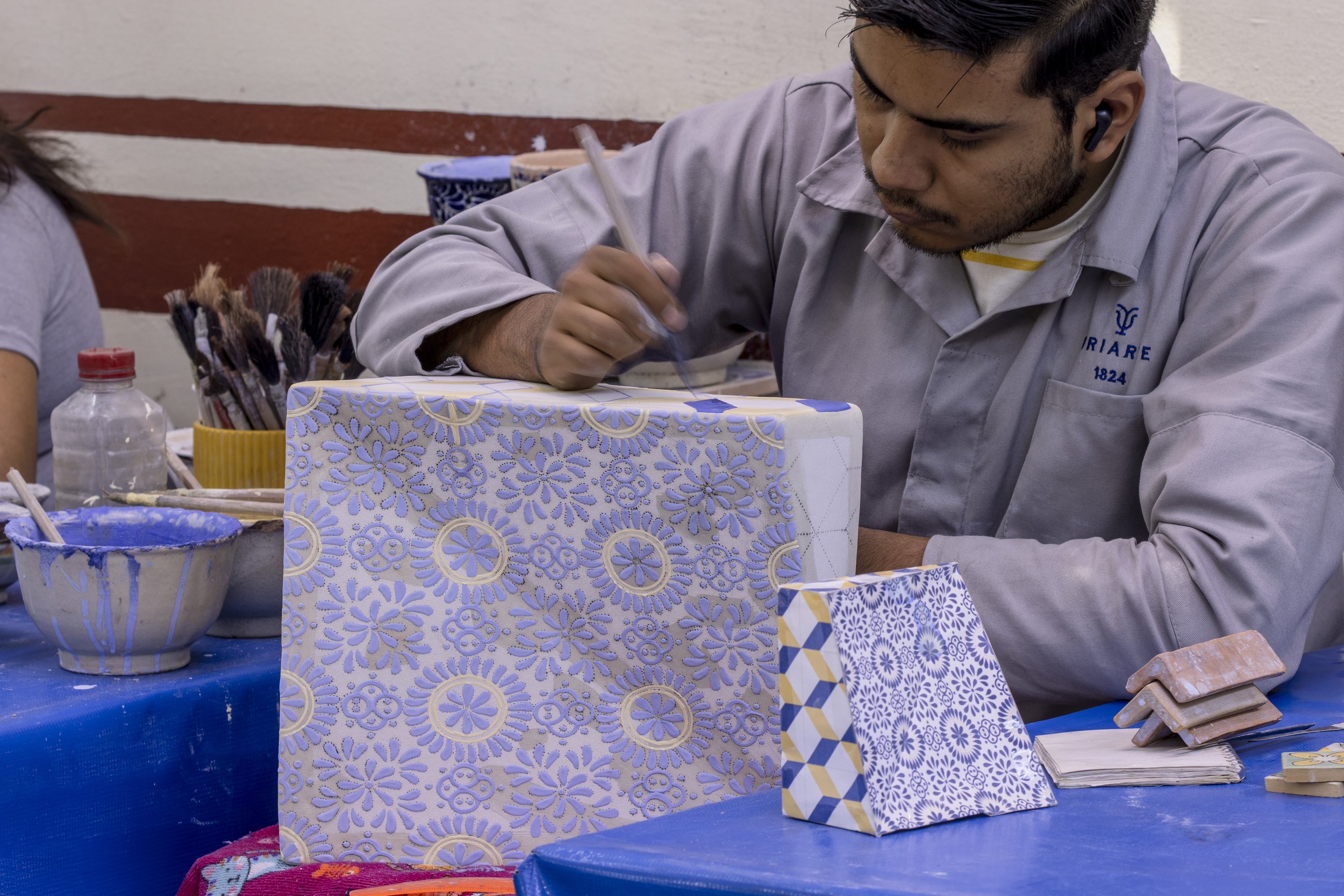 Un hombre decorando un mazo de cartas con patrones azules y dorados en un taller, rodeado de herramientas y materiales de arte.