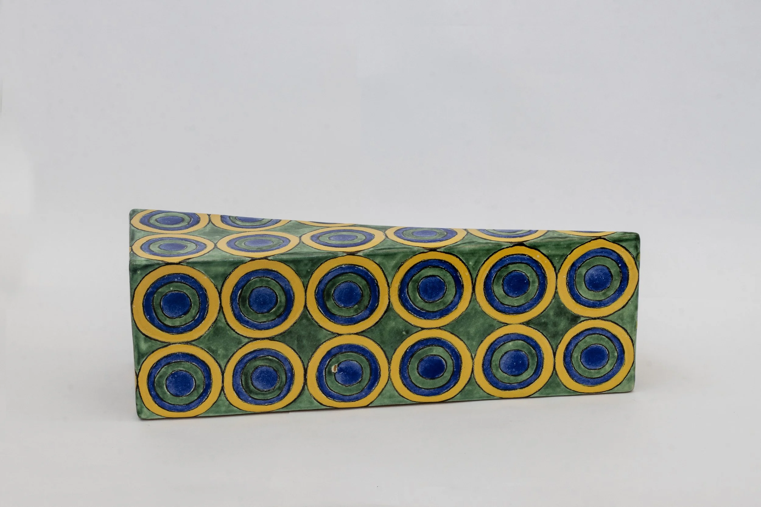 Ladrillo decorativo con patrón de círculos azules y amarillos sobre fondo verde, en un fondo blanco.