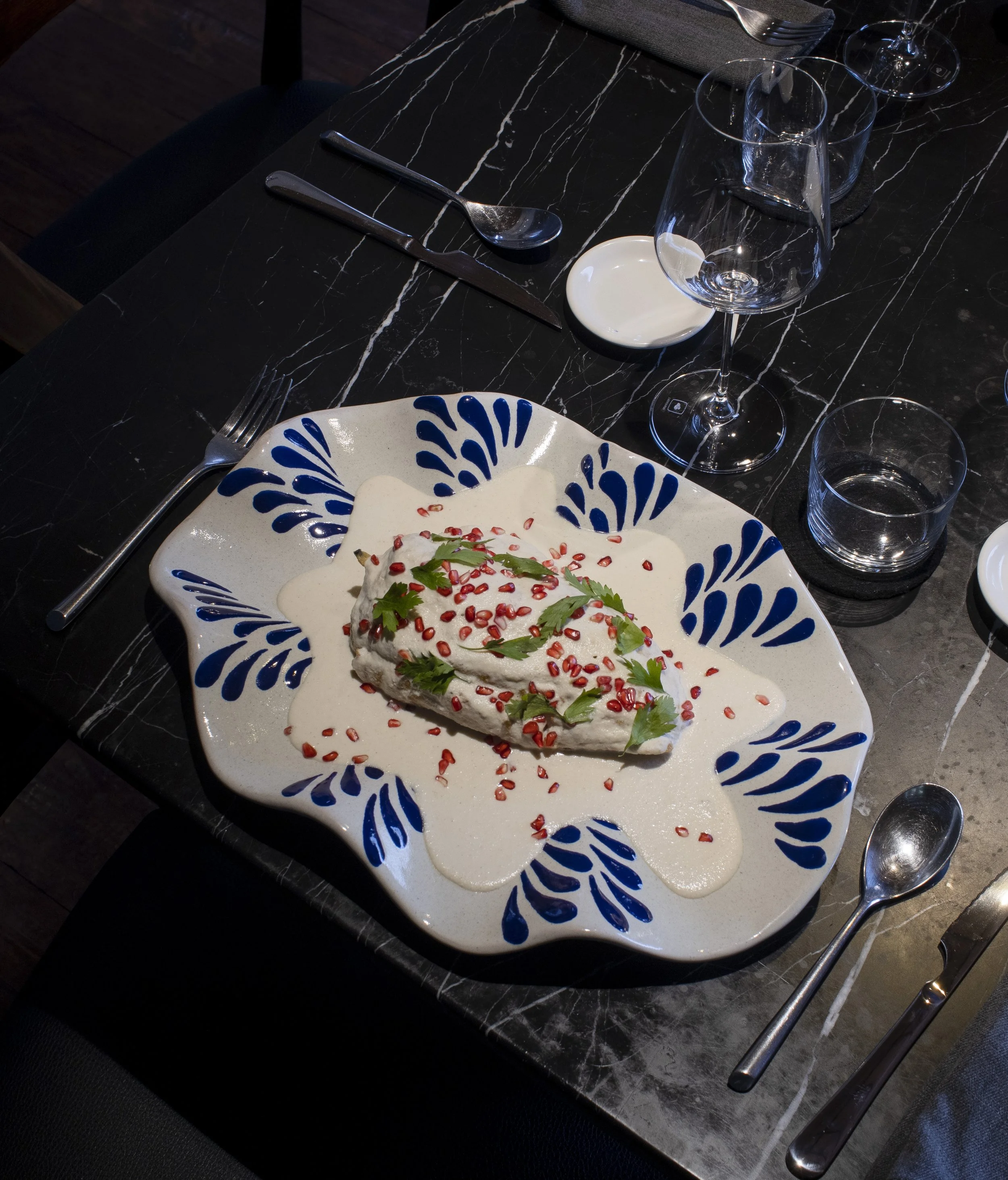 Porción de pescado con salsa blanca, decorada con semillas de granada y perejil, en plato decorativo con borde azul en mesa de mármol negro.