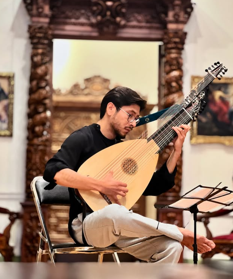 Joven tocando laúd en un salón con decoración clásica, con partituras sobre un soporte y cuadros en las paredes.