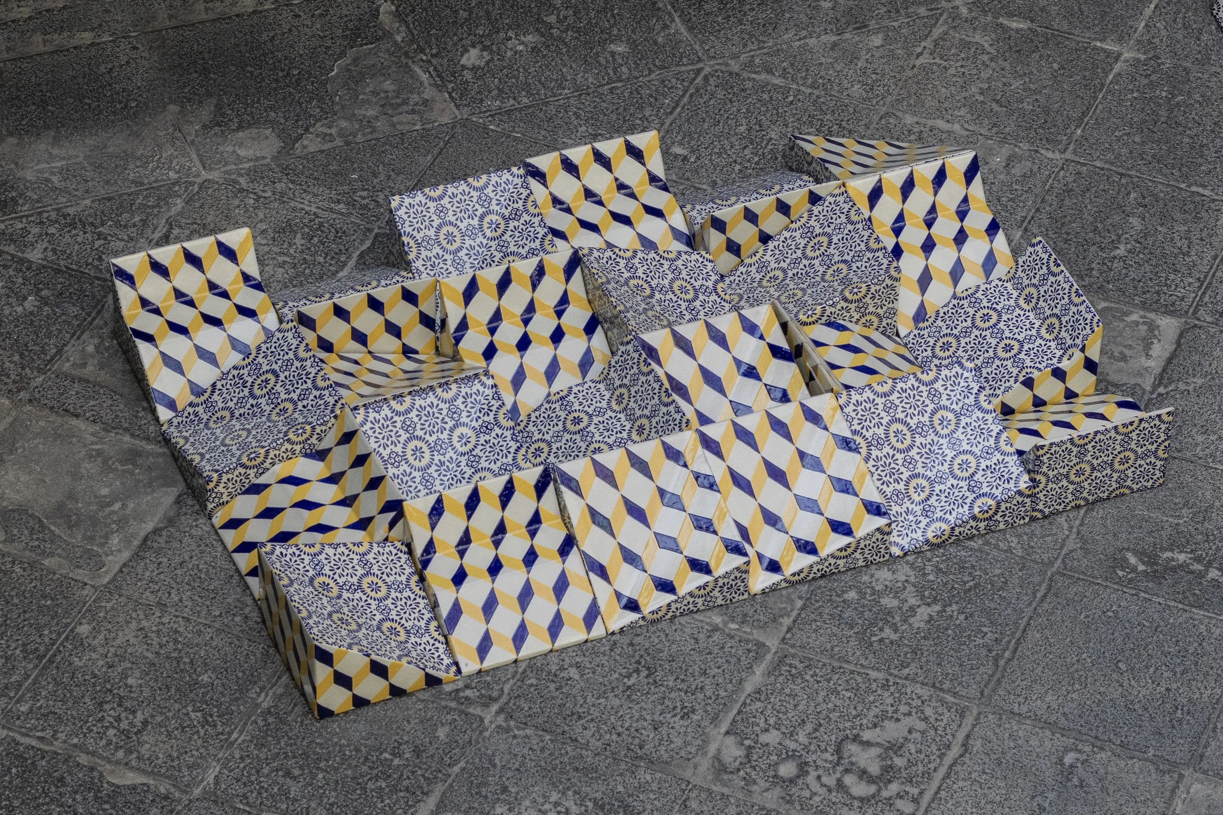 Cubos de cartón decorados con patrones azules, amarillos y blancos, dispuestos sobre un piso de piedra.