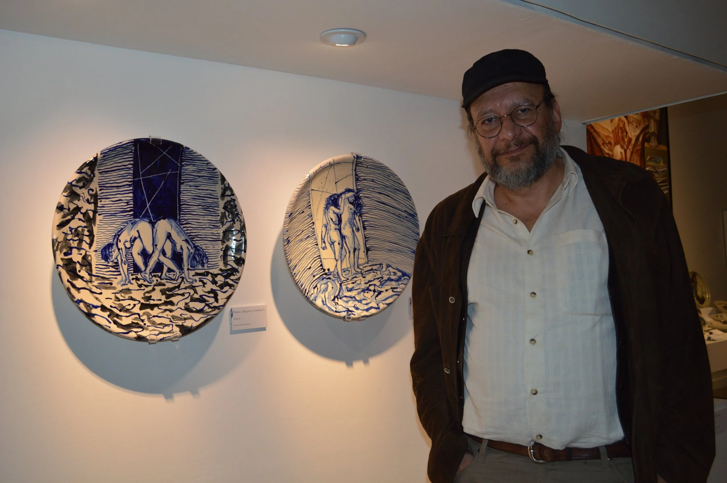 Hombre con barba, lentes y gorra de espaldas en una galería de arte con cerámicas decoradas en azul y blanco