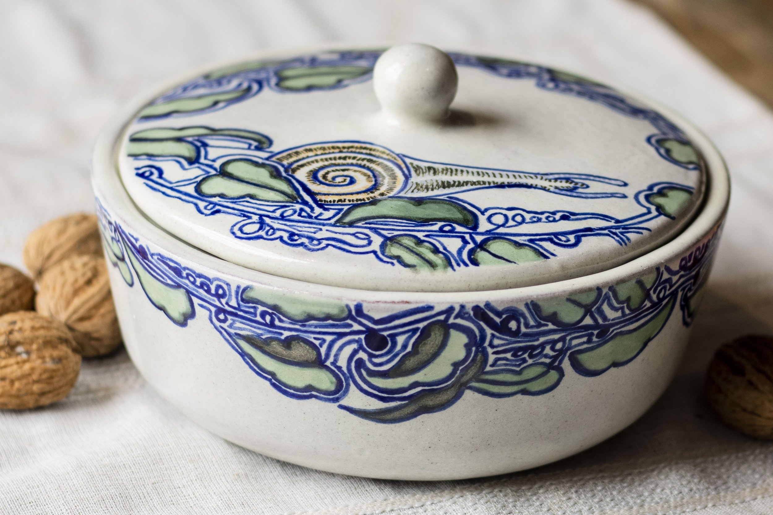 Olla de cerámica decorada con dibujos de líneas azules y verdes, con tapa y pomo, rodeada de nueces.