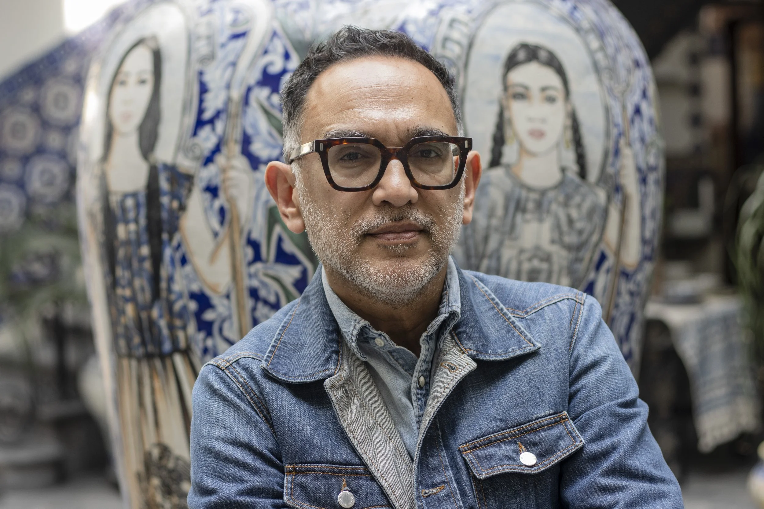 Hombre con gafas de montura gruesa y barba gris, usando chaqueta de mezclilla, en un fondo de azulejos con retratos de mujeres.