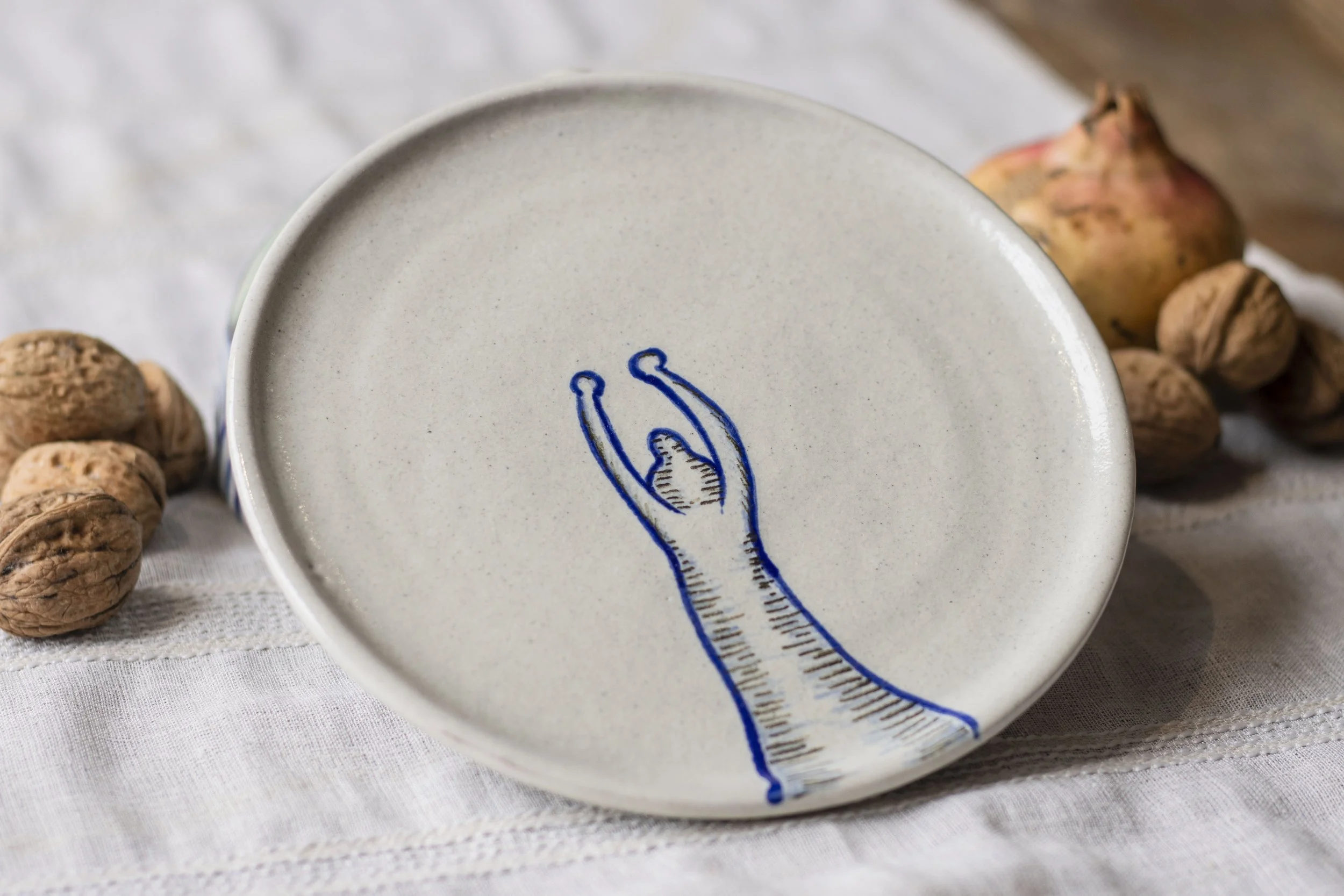 Plato de cerámica con dibujo de una mano en la base, rodeado de nueces y una fruta y nueces en el fondo sobre una tela de lino.