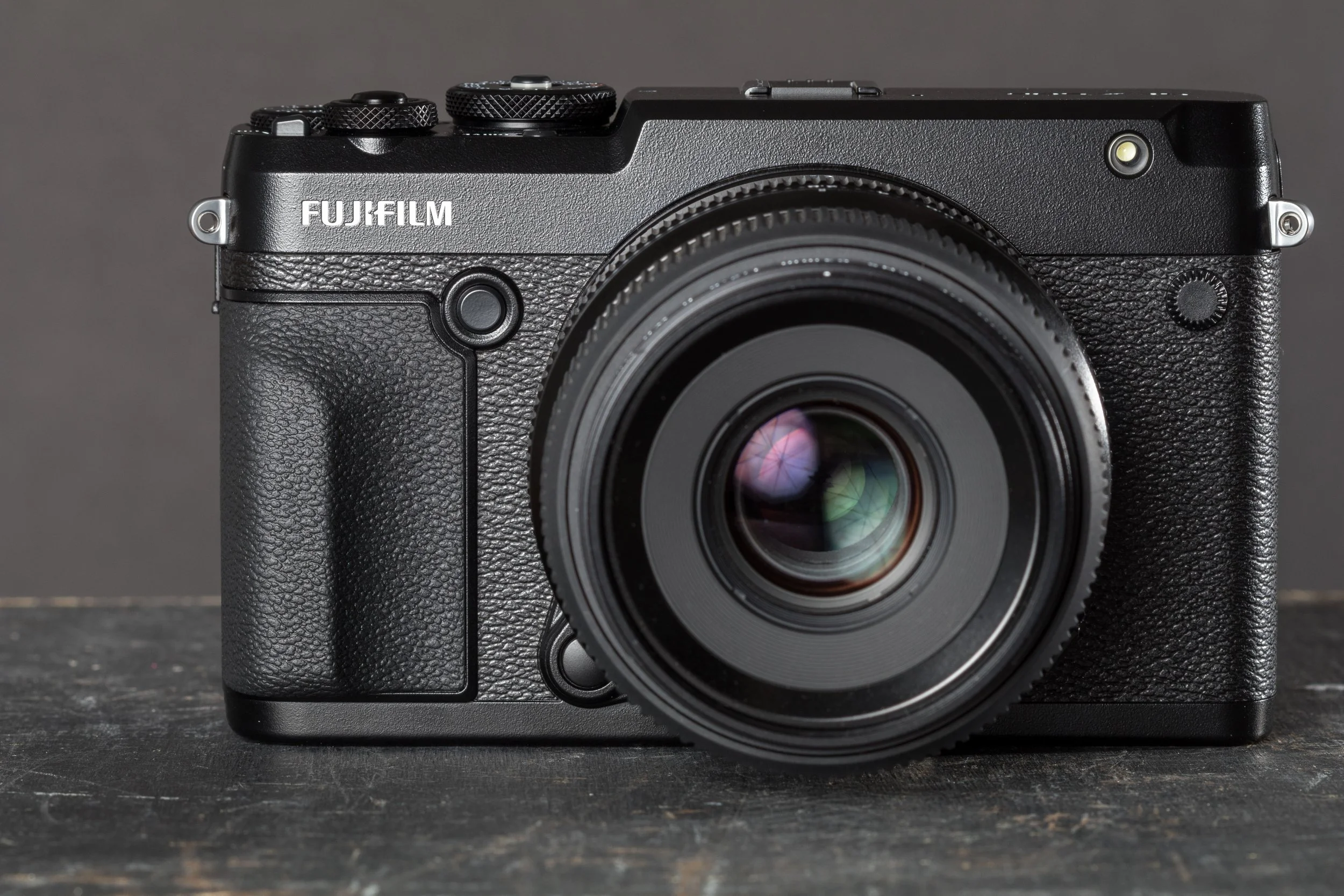 Black Fujifilm digital camera with textured grip and large lens, placed on dark wooden surface against a gray background.