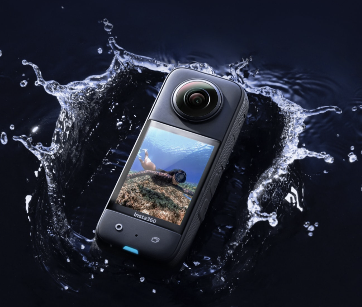 A compact action camera with a large lens, partially submerged in water with splashes around it, displaying an underwater scene on its screen.