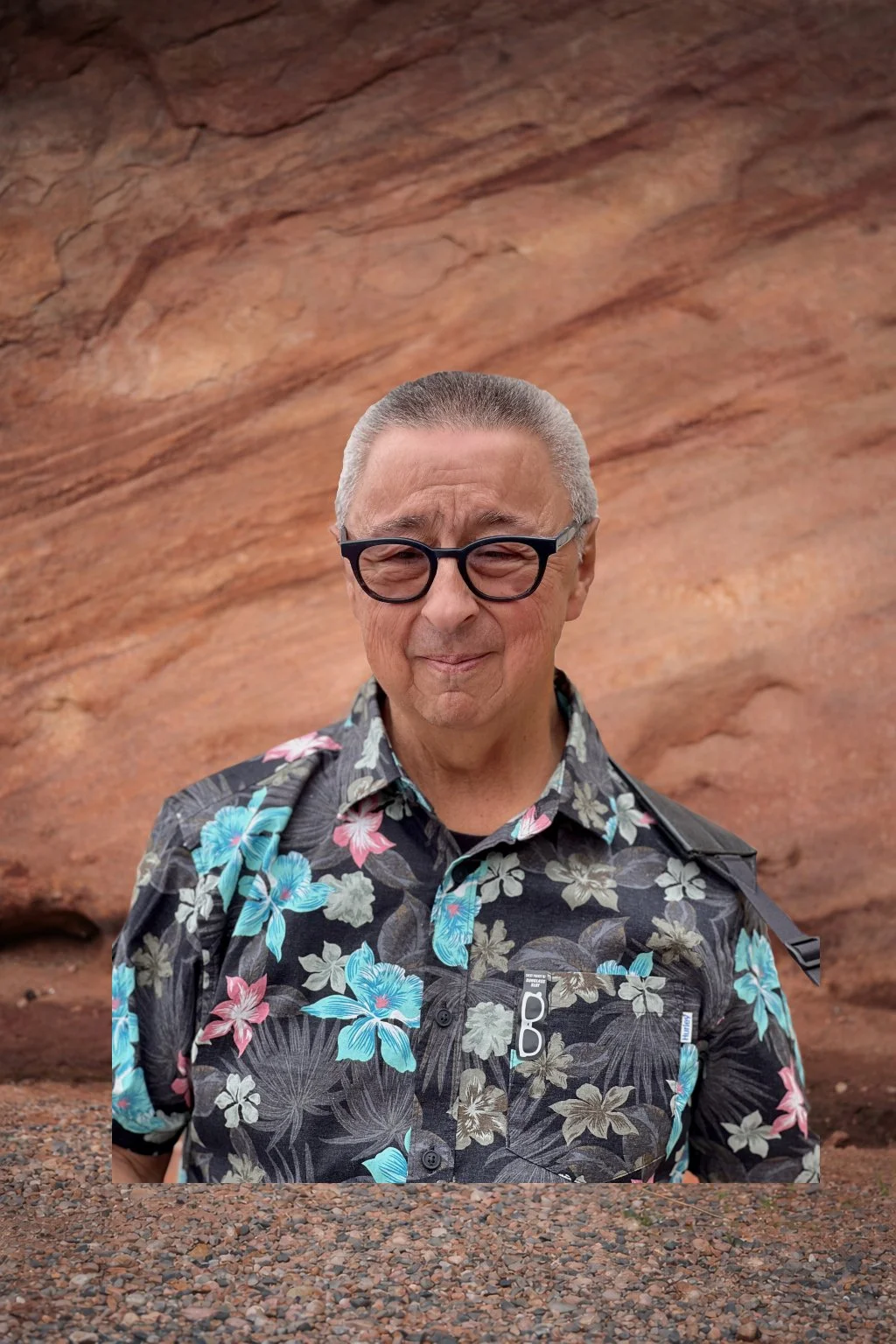 A person wearing glasses and a floral shirt stands in front of a textured brick wall.