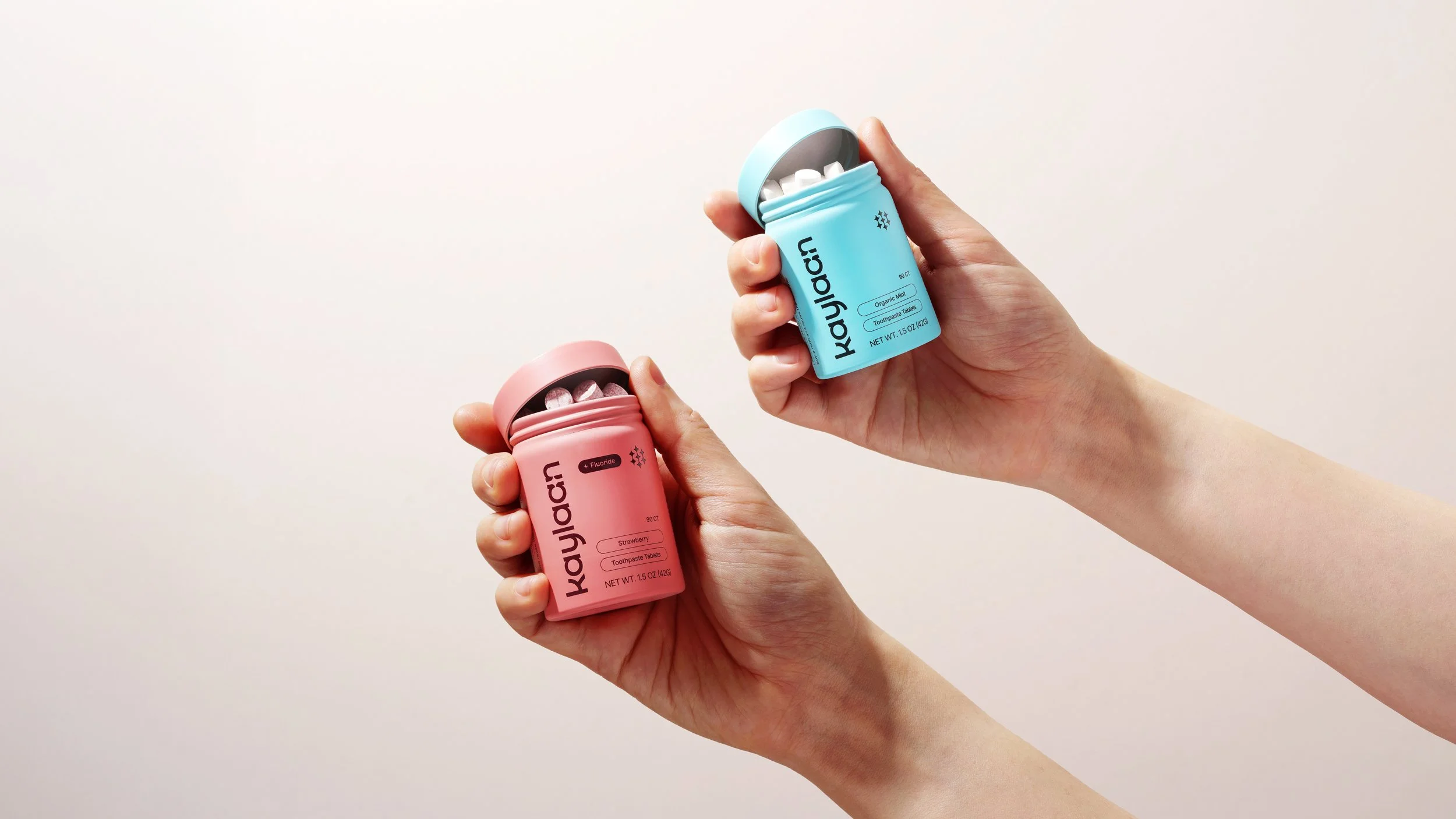Two hands. Each is holding a different flavor of Kaylaan toothpaste tablets for mint and strawberry flavor.