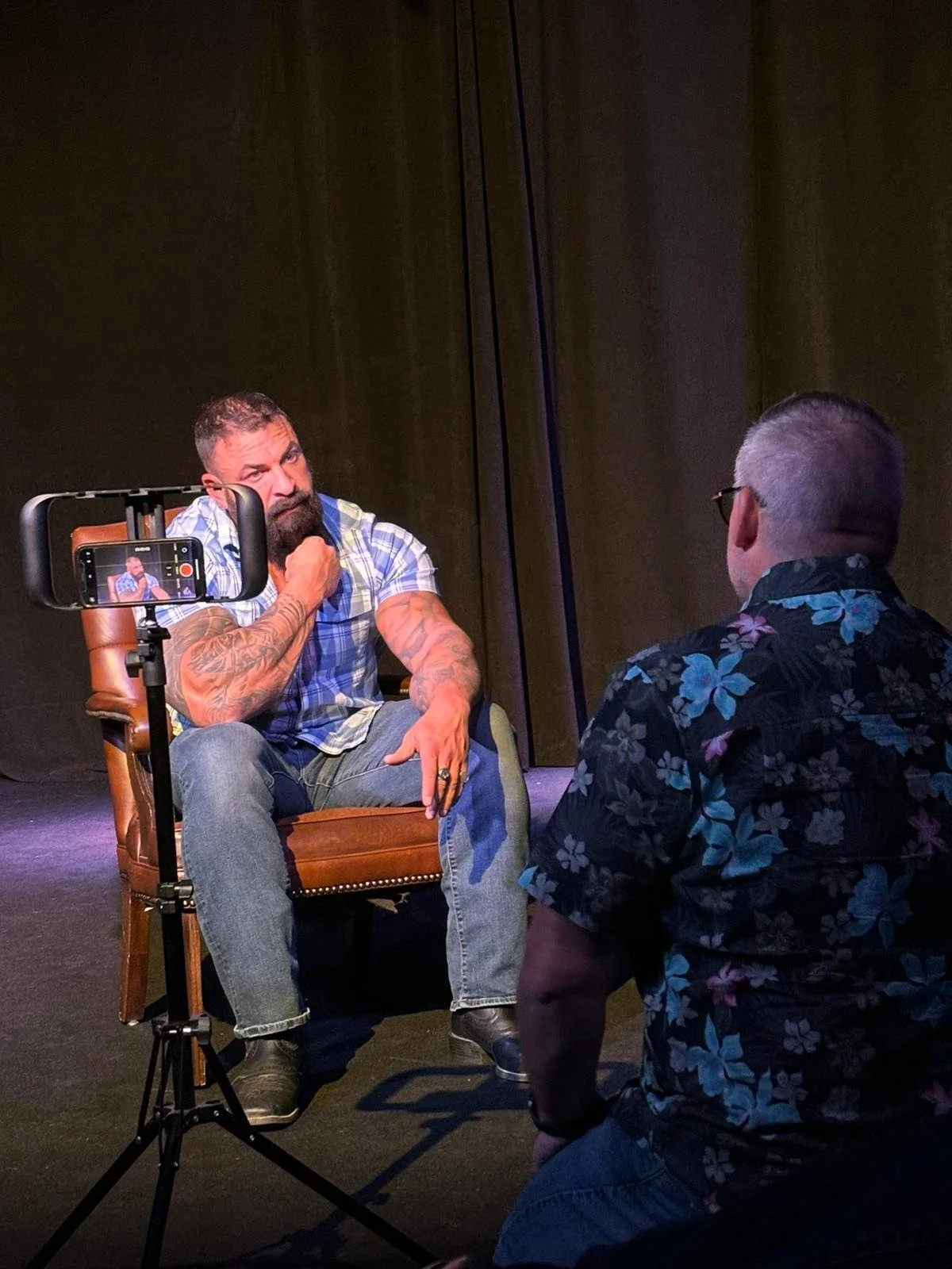 A man with a beard and tattoos sitting in a brown leather chair, being recorded on a smartphone mounted on a tripod, while another man in a floral shirt kneels in front of him talking.